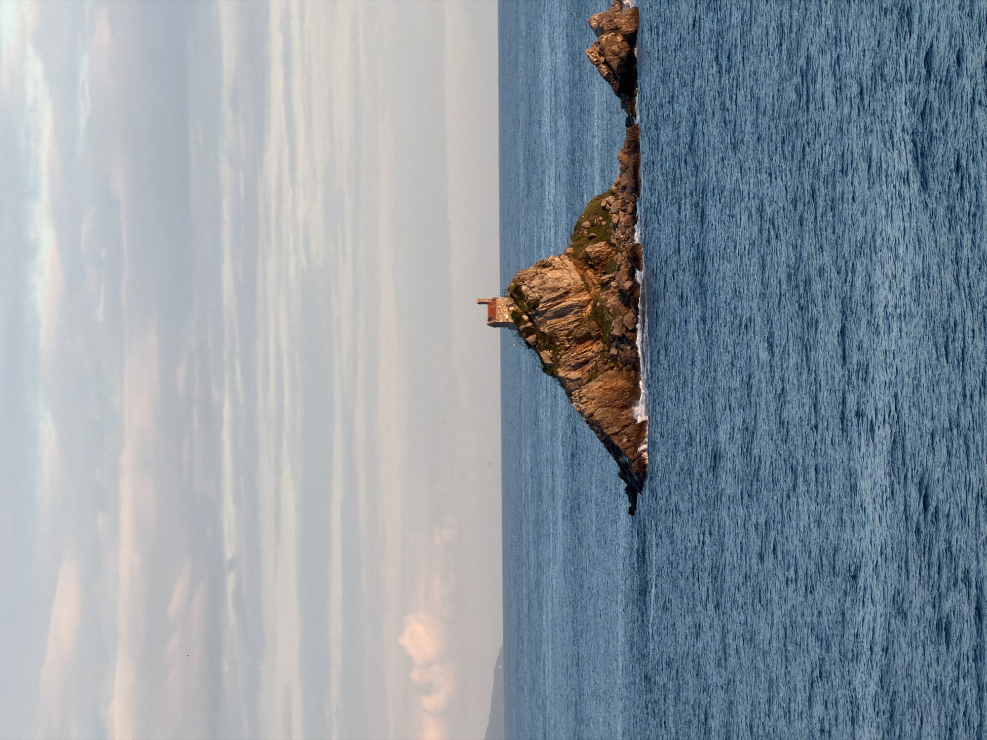 An aerial, rotated perspective of Mirište, Montenegro, featuring a rocky promontory jutting into a body of water on the right and a sky filled with horizontal clouds on the left. The promontory has sparse vegetation and a small structure atop it. The water has ripples visible, suggesting a slight wind.