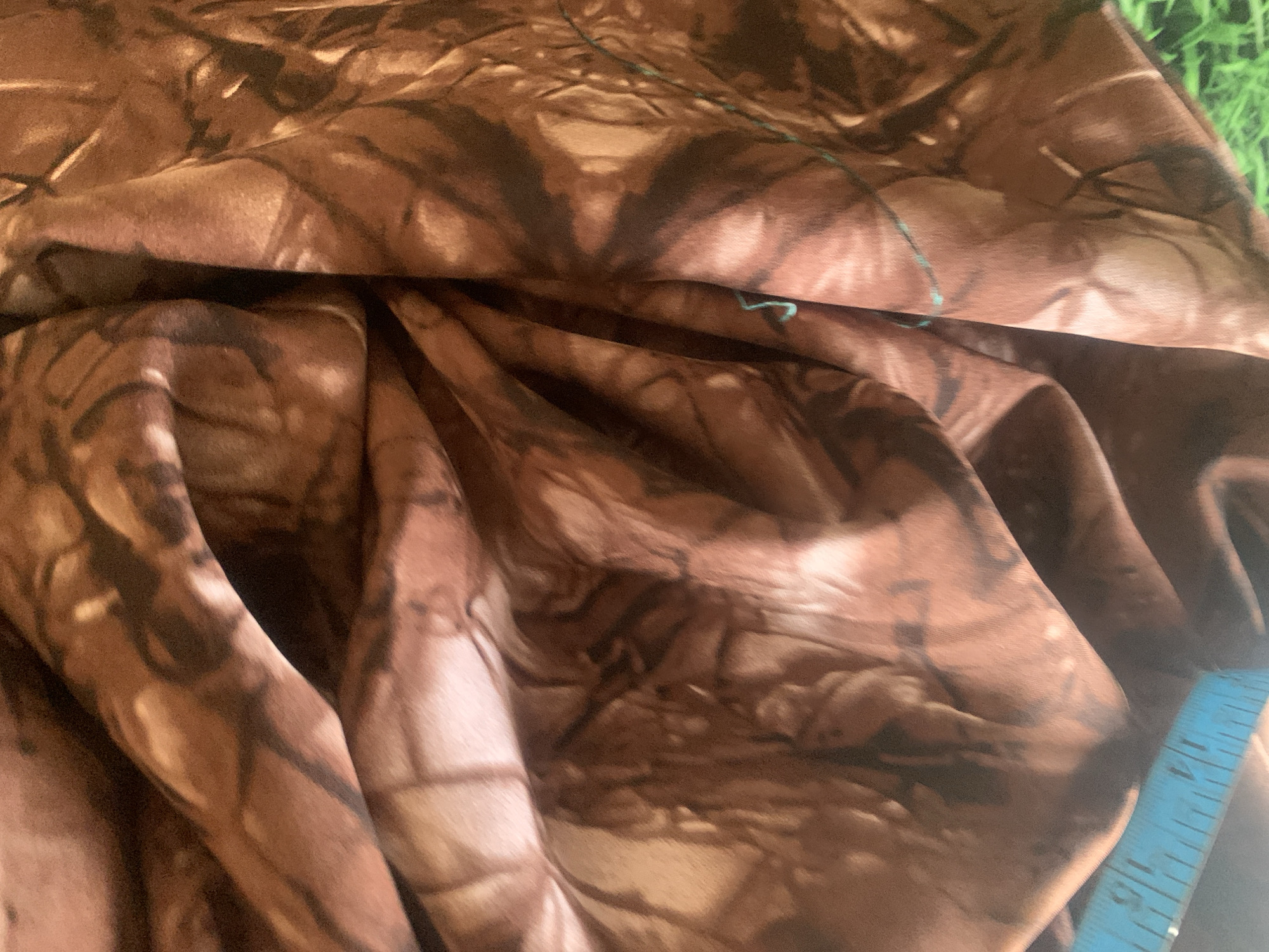 The image displays a close-up view of a brown fabric with a patterned design that resembles tree bark or camouflage. The fabric is draped and folded, creating shadows and highlights that give it a textured appearance. A portion of a blue measuring tape with visible markings is present at the bottom left corner of the image. In the bottom right corner, a glimpse of green grass is visible. The lighting suggests an indoor or shaded outdoor setting. There are no people or animals visible in the image. The scene provides no contextual information related to the city of Jauro Yinu, Nigeria.
