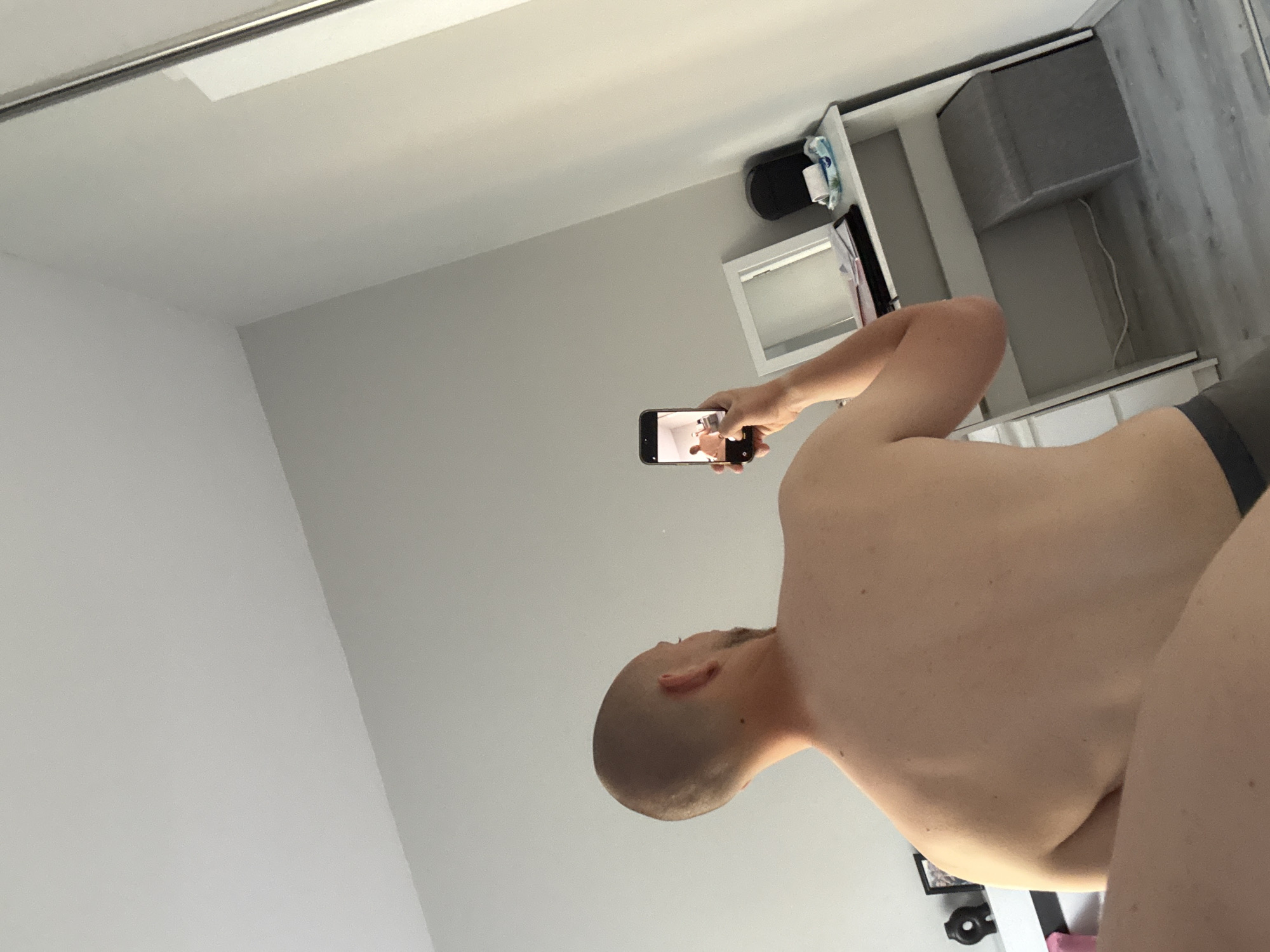A male individual with a shaved head and beard is depicted from behind, shirtless, wearing dark underwear or shorts. He holds a black smartphone in his raised right hand, capturing a selfie in a mirror, as evidenced by the reflection of himself visible on the phone's screen. The scene takes place indoors, in a room with light grey walls and a light grey wooden floor. To the right, a white dressing table or desk is positioned, featuring a rectangular mirror, papers, a black container, and a pack of wipes. A grey storage ottoman or stool is situated beneath the desk, with a white power cord visible on the floor. A partial view of a large mirror's frame is visible on the far right. Objects on a surface, possibly a bedside table, are partially visible on the left.