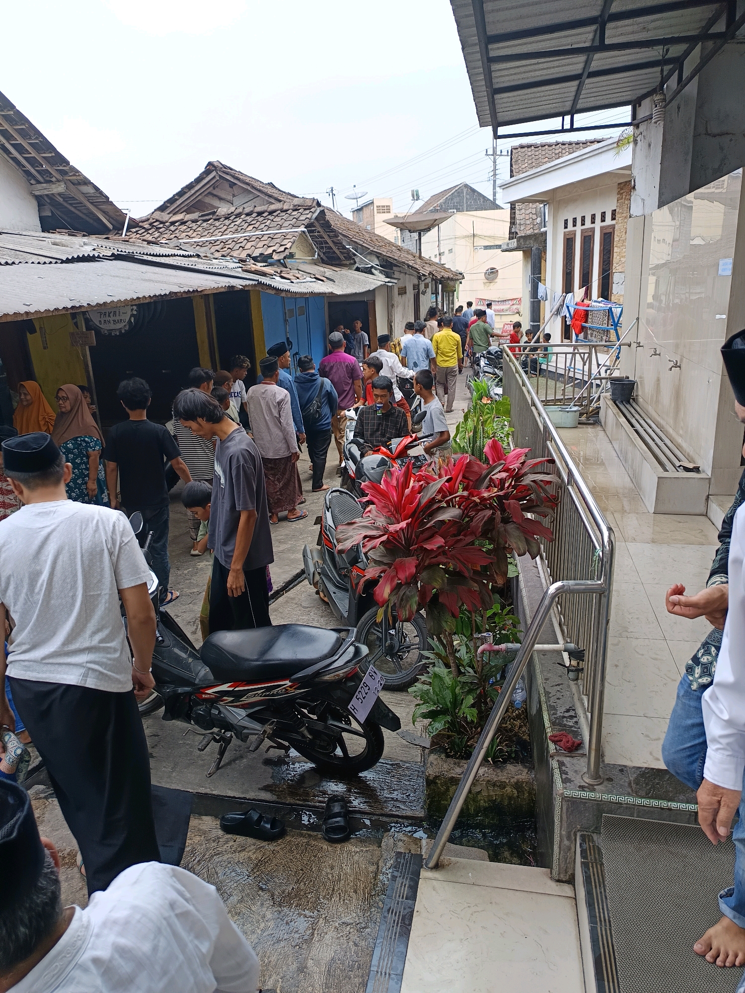 In the city of Klerokrajan, Indonesia, a street scene unfolds with a group of people gathered. The street is narrow and lined with buildings, some appearing to be shops with corrugated metal roofs. Several motorcycles are parked along the pathway, and one is in the foreground, facing left. People are dressed in casual attire, with many men wearing shirts and trousers, and some women in headscarves. The ground is wet and appears to be paved. A metal railing runs along the right side of the image, with plants growing behind it. The overall atmosphere suggests a public gathering or a busy street in a residential or commercial area.