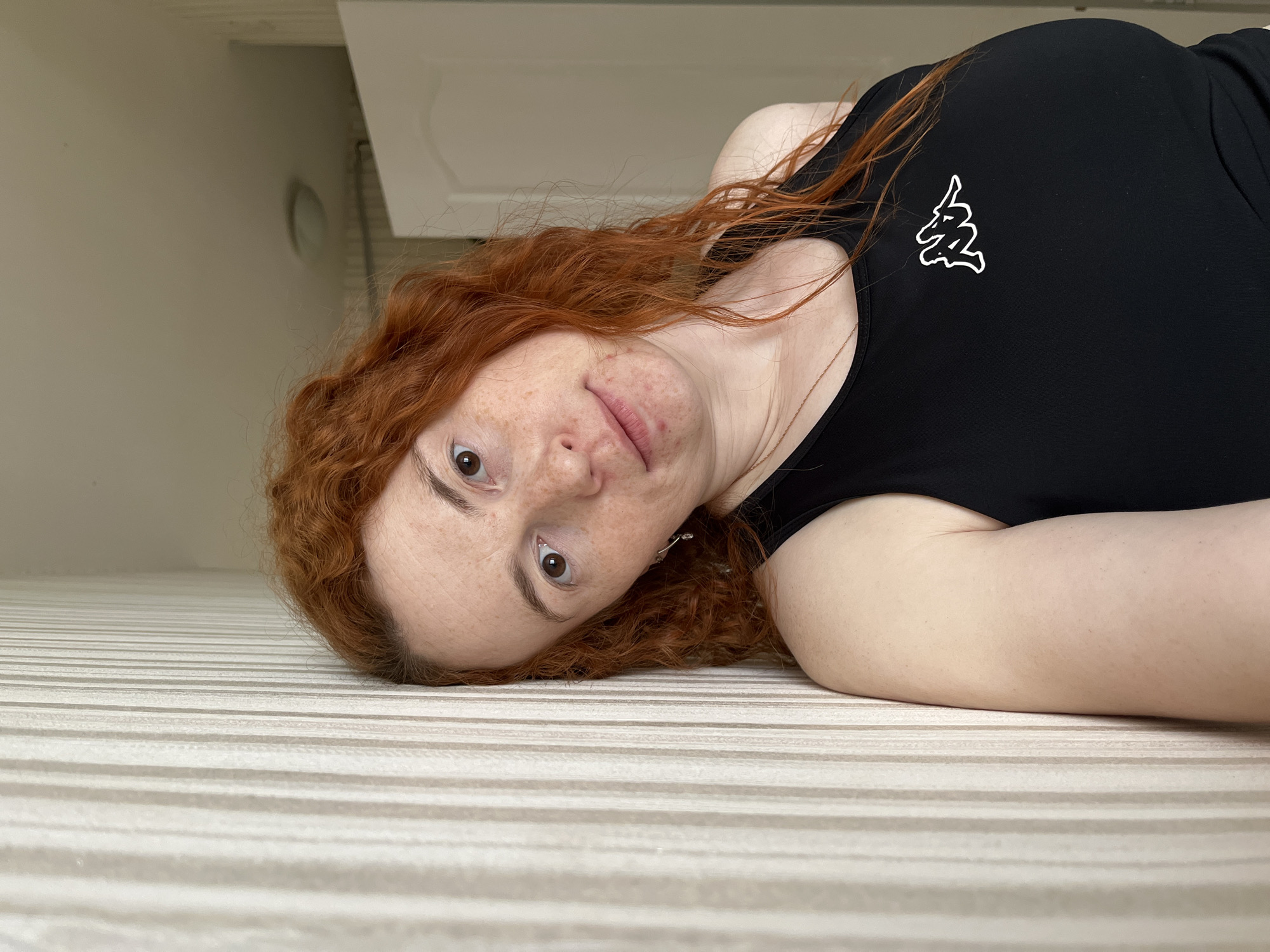 A woman with auburn, curly hair lies on a white and beige striped surface, possibly a bed or a rug. She is wearing a black tank top with a white logo on the upper right. The woman has fair skin with freckles and appears to be looking directly at the camera. A portion of a white wall and a white cabinet or shelf are visible in the background. There is no information present in the provided image indicating the location being the city of Chaikovsky, Russia.