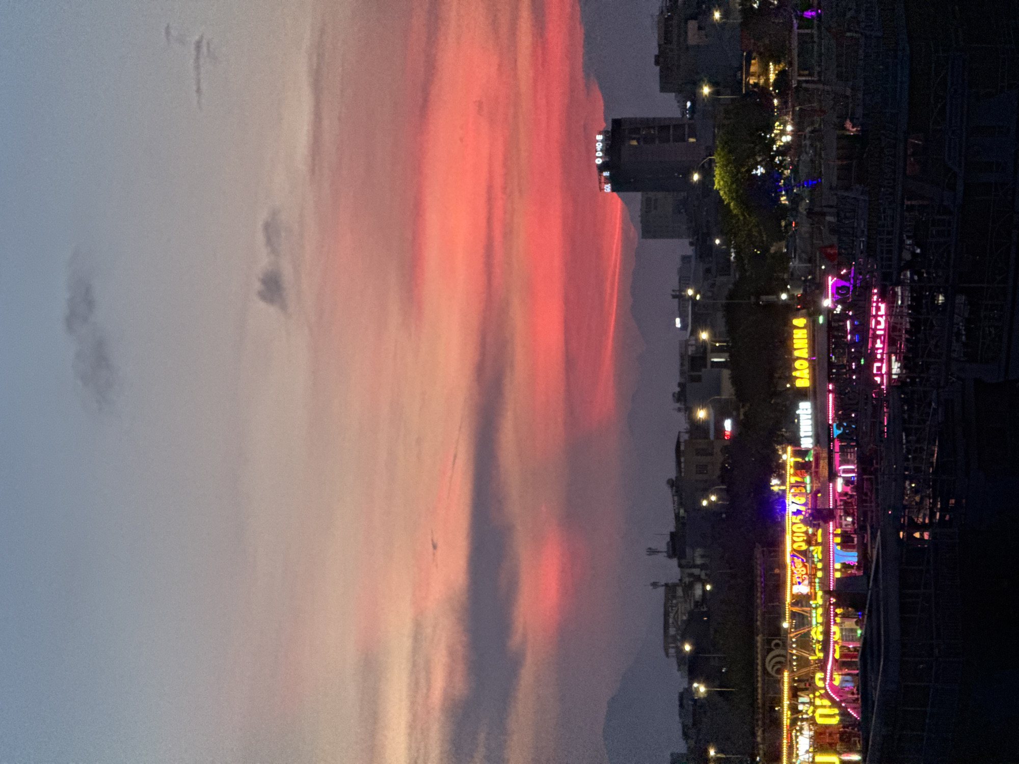The image depicts a sunset over the Han River in Da Nang, Vietnam. The sky is painted with hues of orange, pink, and purple, with scattered clouds. In the foreground, the illuminated boats docked along the riverfront are a vibrant contrast to the darkening sky. These boats are adorned with numerous colorful lights, primarily in yellow, pink, and blue, creating a festive and lively atmosphere. One boat has signage that reads "BAO ANH 4" in yellow neon letters. In the background, the city skyline is visible, with buildings and streetlights, and a tall building with illuminated signage that reads "GOLDEN MOON" is prominent. Mountains form a silhouette against the horizon. The overall scene captures the bustling nightlife of Da Nang along its river.