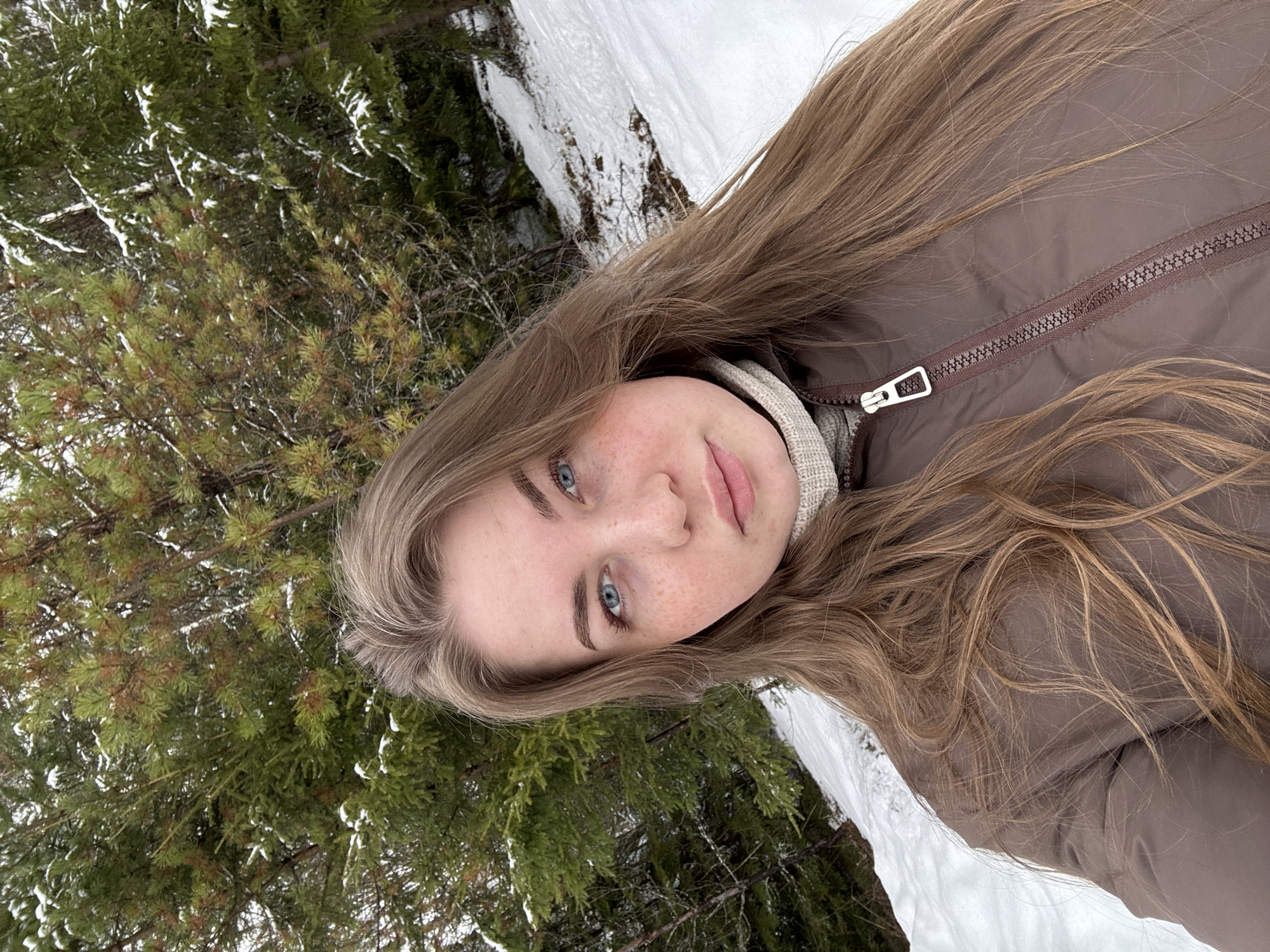 The image is a selfie taken outdoors. The subject is a fair-skinned young woman with light brown, long hair. She has blue eyes and freckles. She is wearing a beige sweater and a brown jacket that is zipped up. The background consists of evergreen trees with some snow on the branches, and a snow-covered ground area. The environment suggests a forest setting. The location is Zavod Mikhailovskii, Russia.