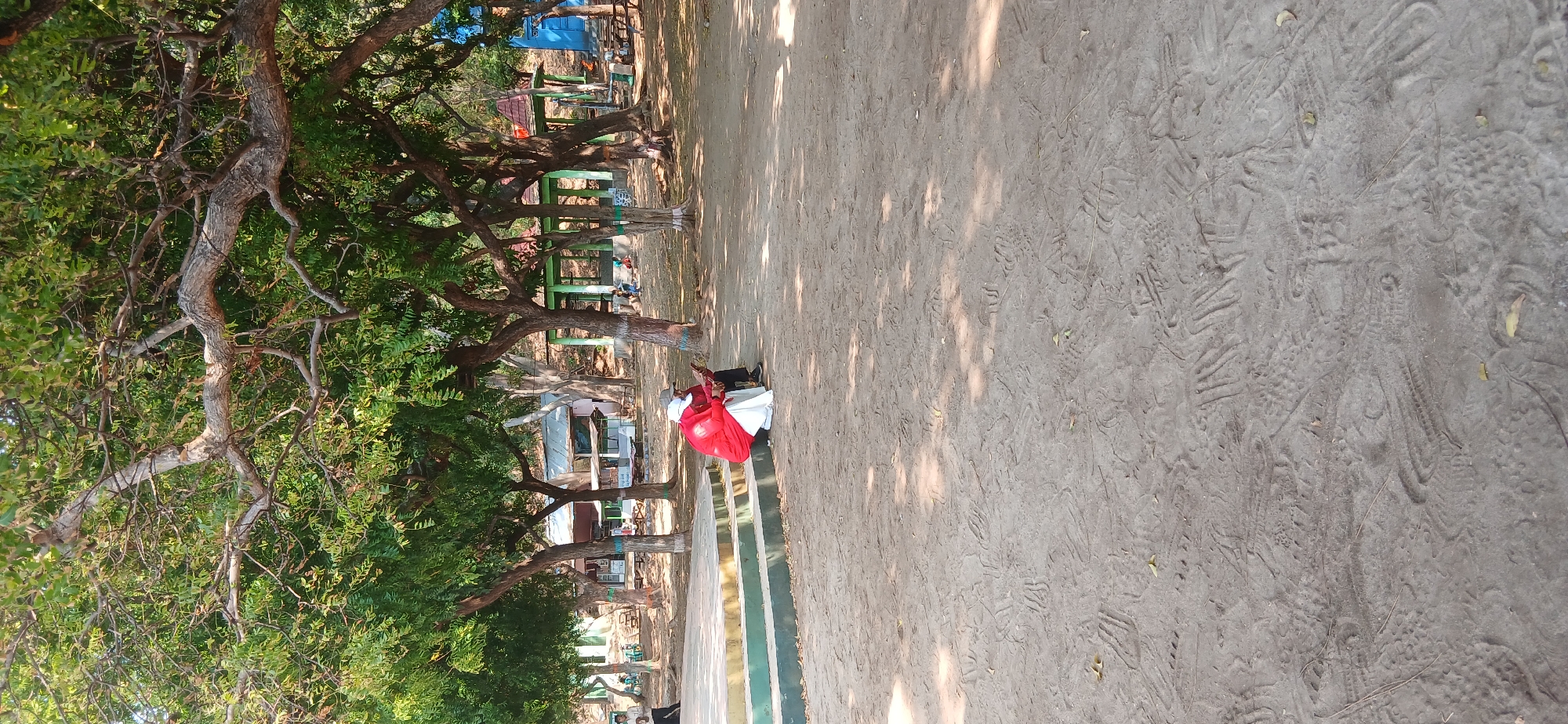 An outdoor scene in Banyuglugur, Indonesia, features a sandy ground with numerous footprints. In the middle ground, a person wearing a red shirt and white head covering sits on the edge of a tiered platform, looking down at a device in their hands. Behind them, several trees with leafy canopies provide shade, and beyond the trees are buildings, some with peaked roofs, and additional trees. To the far left, a person stands near the platform. The overall environment suggests a park or public gathering space.