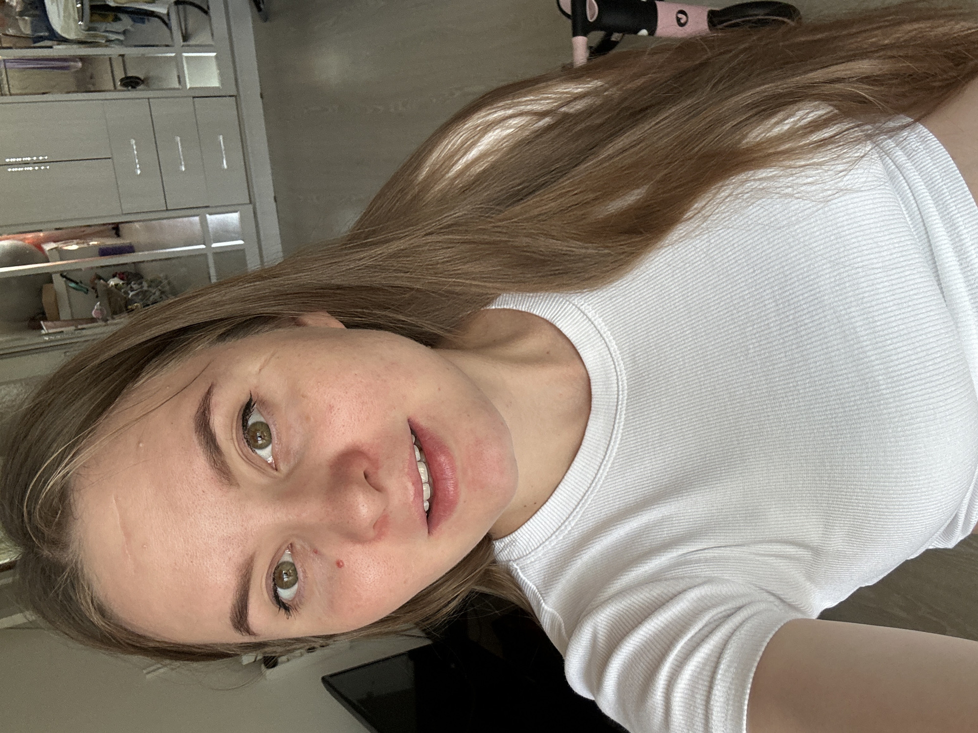The image shows a close-up, selfie-style shot of a young woman with long, light brown hair. She is wearing a white ribbed t-shirt and appears to be indoors. Her facial features include green eyes, light skin with some blemishes, and a light pinkish tone around her nose and cheeks. She is looking directly at the camera with a slight smile. In the background, a white wardrobe with mirrored doors is visible, along with a television screen on the left. A pink and black child's bicycle is partially visible on the right side of the frame. The floor appears to be light-colored wood or laminate. The scene is likely a residential interior. There is no information about the location being in the city of Noviy, Russia.
