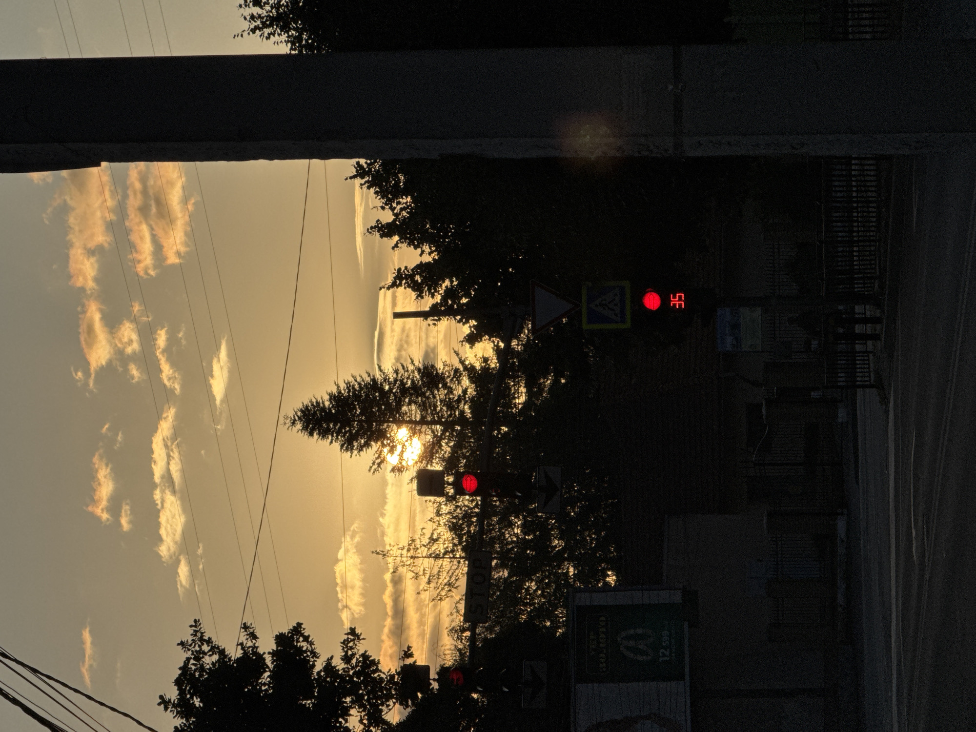 The image captures a street scene at sunset in Chernivtsi, Ukraine. The sky is a warm gradient of orange and yellow, with scattered clouds catching the last light. Silhouetted against the sky are trees and utility wires. In the foreground, a traffic light displays a red signal, accompanied by a speed limit sign showing "35". To the left, another traffic light is also red, and a "STOP" sign is visible. A pedestrian crossing sign is also present. The overall impression is that of an urban intersection at dusk, with the sun setting behind the trees, casting long shadows and creating a dramatic silhouette effect.