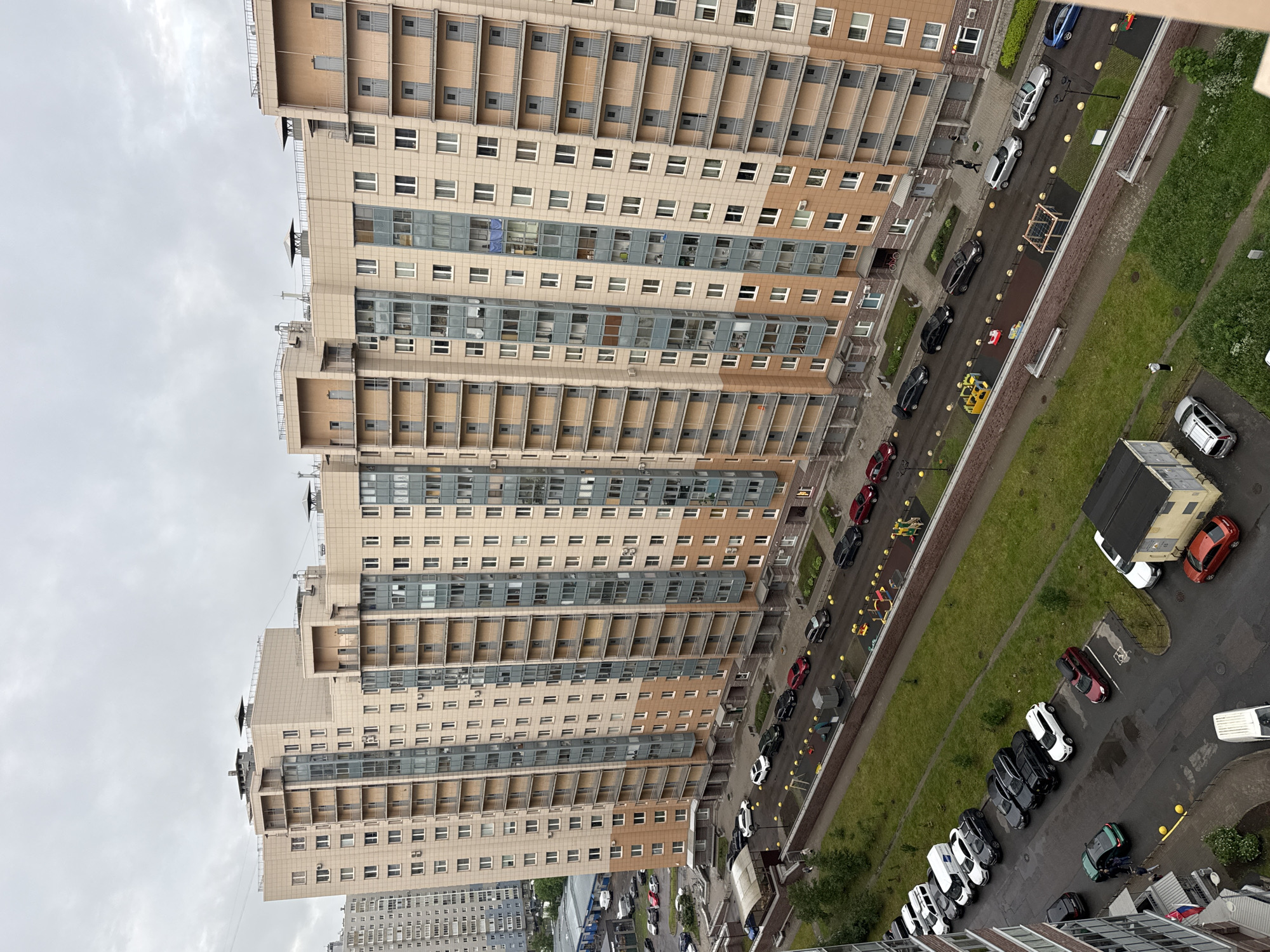 The image shows a view from a high vantage point overlooking a residential area with large, modern apartment buildings. The sky is overcast, indicating a cloudy day. In the foreground and midground, several cars are parked along a paved street and in designated parking areas. The apartment buildings are tall and feature a consistent architectural style with balconies, numerous windows, and a light-colored facade with some brown accent sections. There are also glimpses of playgrounds and green grassy areas between the buildings and the parking lots. On the far left, a slightly older, multi-story building with a lighter, more uniform facade is visible. In the lower portion of the image, there are a few individuals walking on the sidewalk and near parked vehicles. The scene appears to be a typical urban or suburban residential complex.