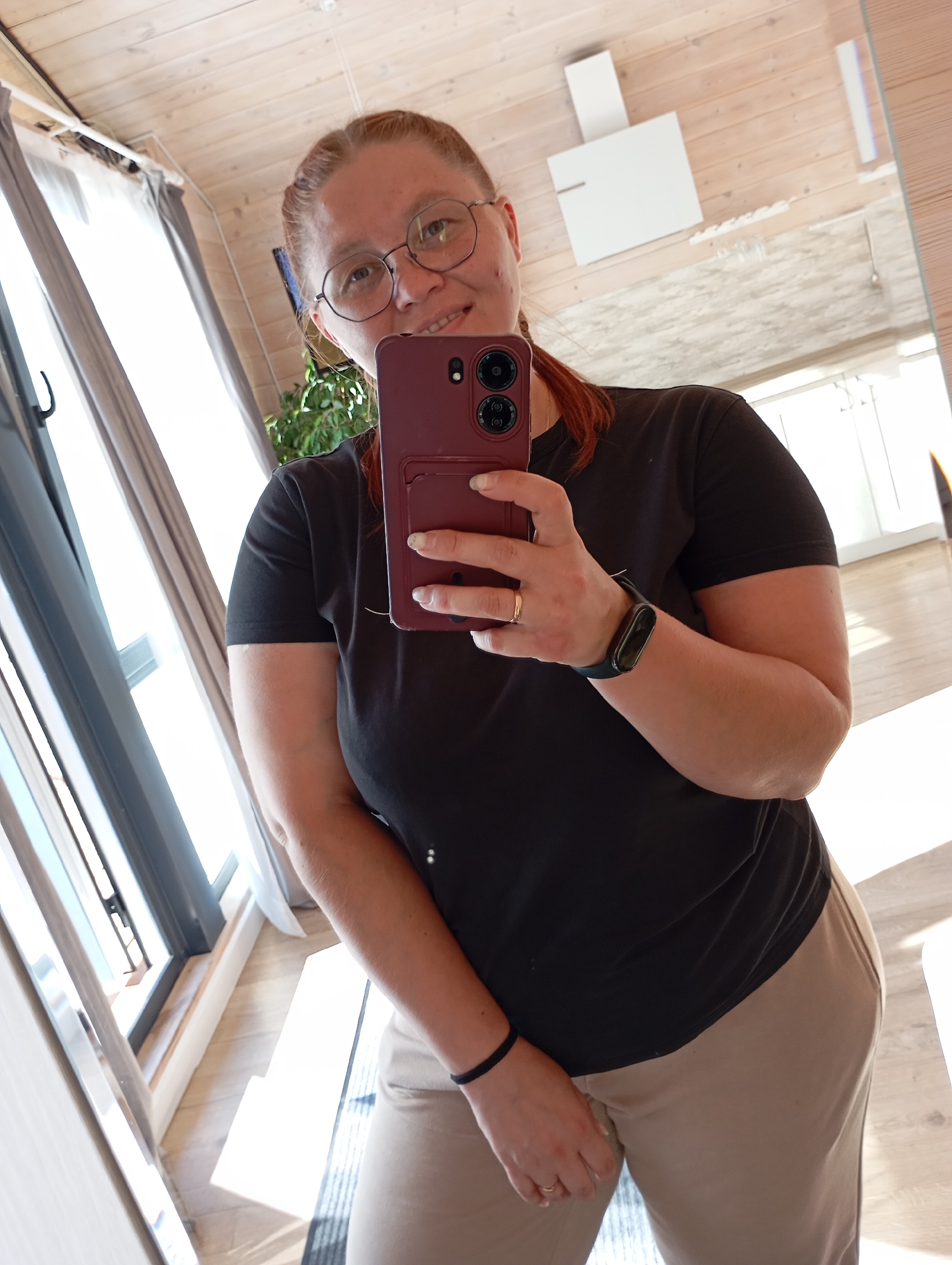A woman with light brown hair, wearing glasses and a black t-shirt, takes a mirror selfie in what appears to be a modern apartment. She is holding a burgundy smartphone with a camera bump. A black fitness tracker is visible on her left wrist, and a thin gold ring is on her left ring finger. She is also wearing tan pants and a black bracelet on her right wrist. The background shows a ceiling with wooden planks, a large window with grey curtains, and some furniture reflected in a mirror. The floor is made of light-colored wood planks. The image is well-lit, with sunlight streaming in from the window. The scene is likely indoors, in a residential setting. There is no indication that this location is in the city of Malyi Bukor, Russia.