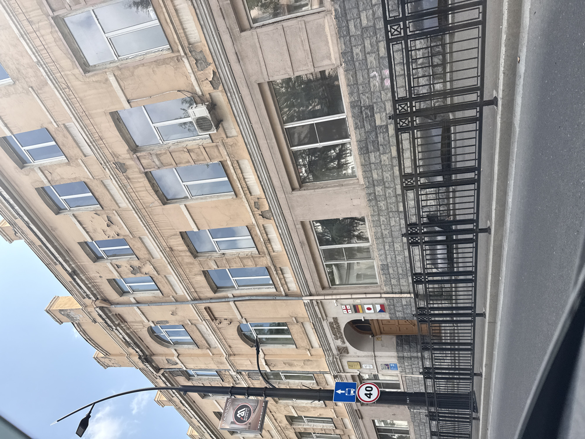 The image shows the facade of a building in Tbilisi, Georgia, likely a school or administrative building, with a prominent arched entryway. The building features several floors with numerous windows, many of which have white frames. The exterior walls are painted a light peach color and show signs of wear and peeling paint, particularly along the upper cornices and around the window frames. The lower portion of the building is clad in grey stone blocks.
In front of the building, a black metal fence with decorative elements lines the sidewalk. A street lamp with a modern design stands to the left of the frame. A sign on the lamp post displays text in Georgian and English, indicating a business or institution. Below this, a blue traffic sign with a white arrow points upwards, and a circular speed limit sign displays "40" within a red border.
The entryway features a wooden door, and above it, several flags are displayed: the flag of Georgia, the flag of Germany, the flag of Japan, and the flag of the Czech Republic. This suggests a connection to international relations or a diverse student body.
The image is taken from street level, looking slightly upwards at the building. The street in the foreground is paved, and a sidewalk separates it from the fence and building. The sky above is blue with some light clouds. The overall impression is of an older urban setting in Tbilisi.