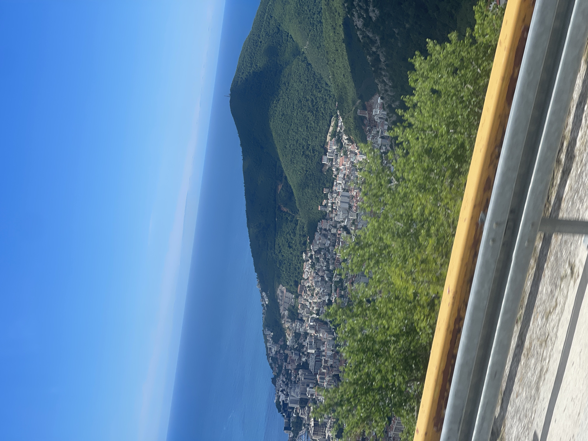 The image captures a view of the city of Budva, Montenegro, from an elevated perspective, likely a viewpoint along a road or highway. The scene includes a blue sky and sea, a mountain densely covered with green trees, and a cluster of buildings that make up the city. A metal guardrail, consisting of yellow and silver horizontal bars, is visible in the foreground. A tree is partially visible.