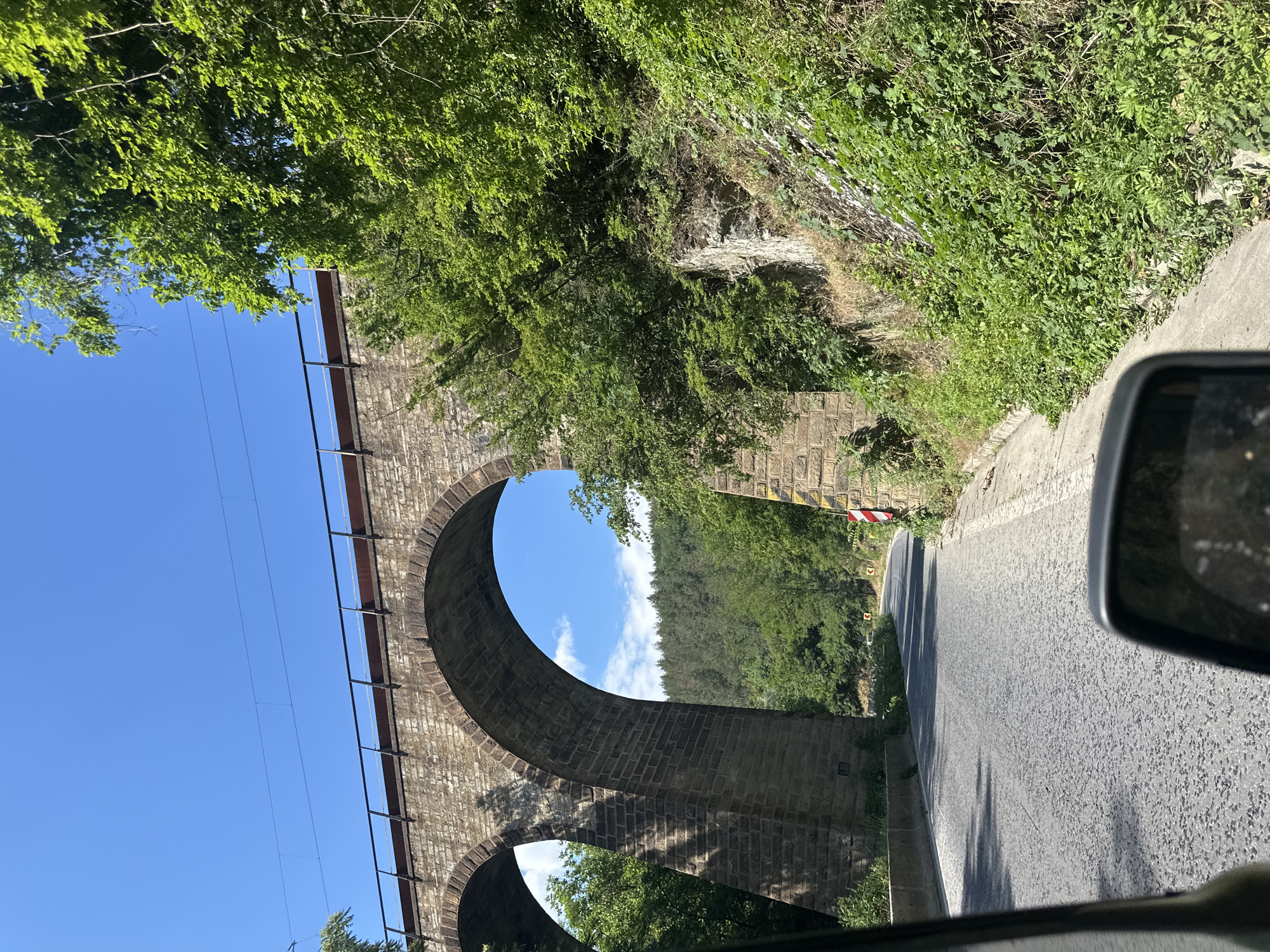 The image depicts a road passing under a large, stone arched viaduct in Zmeyovo, Bulgaria. The viaduct, constructed from rough-hewn, beige-colored stones, features at least two prominent arches, with the closest arch dominating the left side of the frame. The top of the viaduct is lined with a metal railing, and what appear to be train tracks or overhead wires are visible against the clear blue sky.
The road itself is paved with asphalt and shows signs of wear. It curves gently to the right, disappearing into the distance. The sides of the road are lined with dense, green vegetation, including trees and bushes, particularly on the right embankment where rocky outcrops are visible. A red and white striped marker pole stands on the right side of the road, indicating a potential hazard or boundary.
In the foreground, the side mirror of a vehicle is visible in the lower right corner of the image, suggesting the photo was taken from inside a car. The lighting indicates it is daytime, with sunlight casting shadows. The background reveals a forested hillside under a bright blue sky with scattered white clouds. No people or animals are present in the image.
