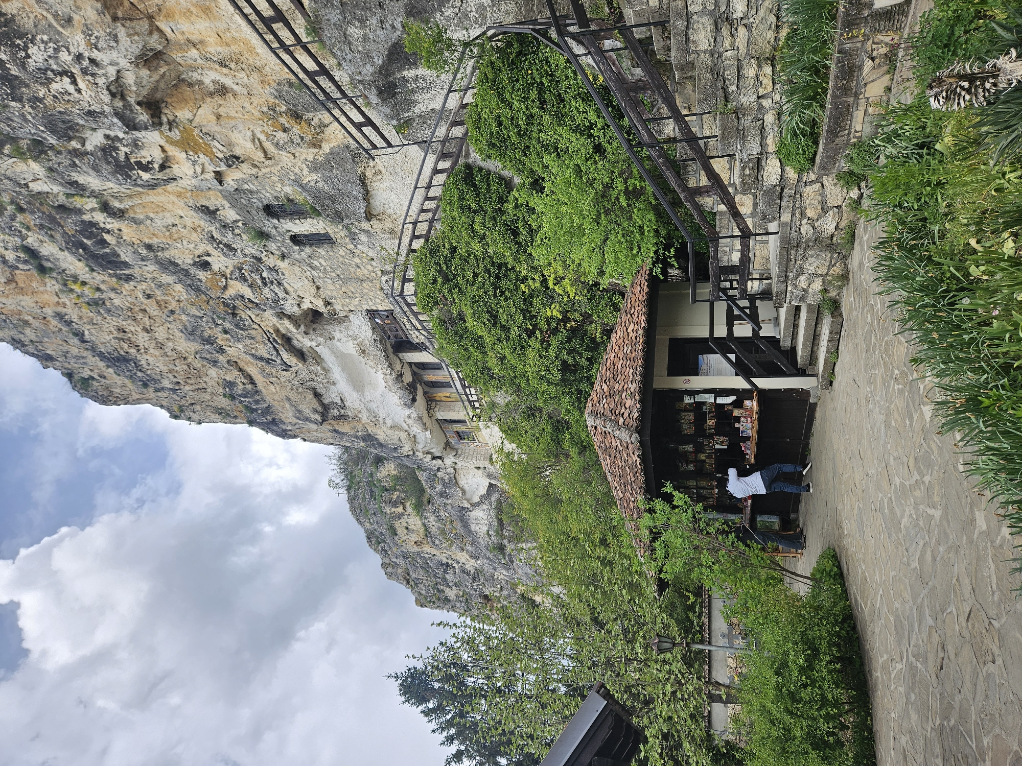 The image captures a scene at the Rock Monastery of Saint Dimitar Basarbowski, located near Basarbovo, Bulgaria. A large, weathered cliff face dominates the left side of the frame. Buildings, likely part of the monastery, are built into the cliff face. A metal walkway and staircase are visible along the cliff.
A small shop with a tiled roof is situated at the base of the cliff. Inside the shop, a person wearing a white shirt is standing behind a counter. The shop appears to sell souvenirs or religious items. A stone-paved path leads towards the shop. Lush green vegetation grows around the buildings and path.
The sky with white, grey, and blue clouds occupies the top portion of the image.