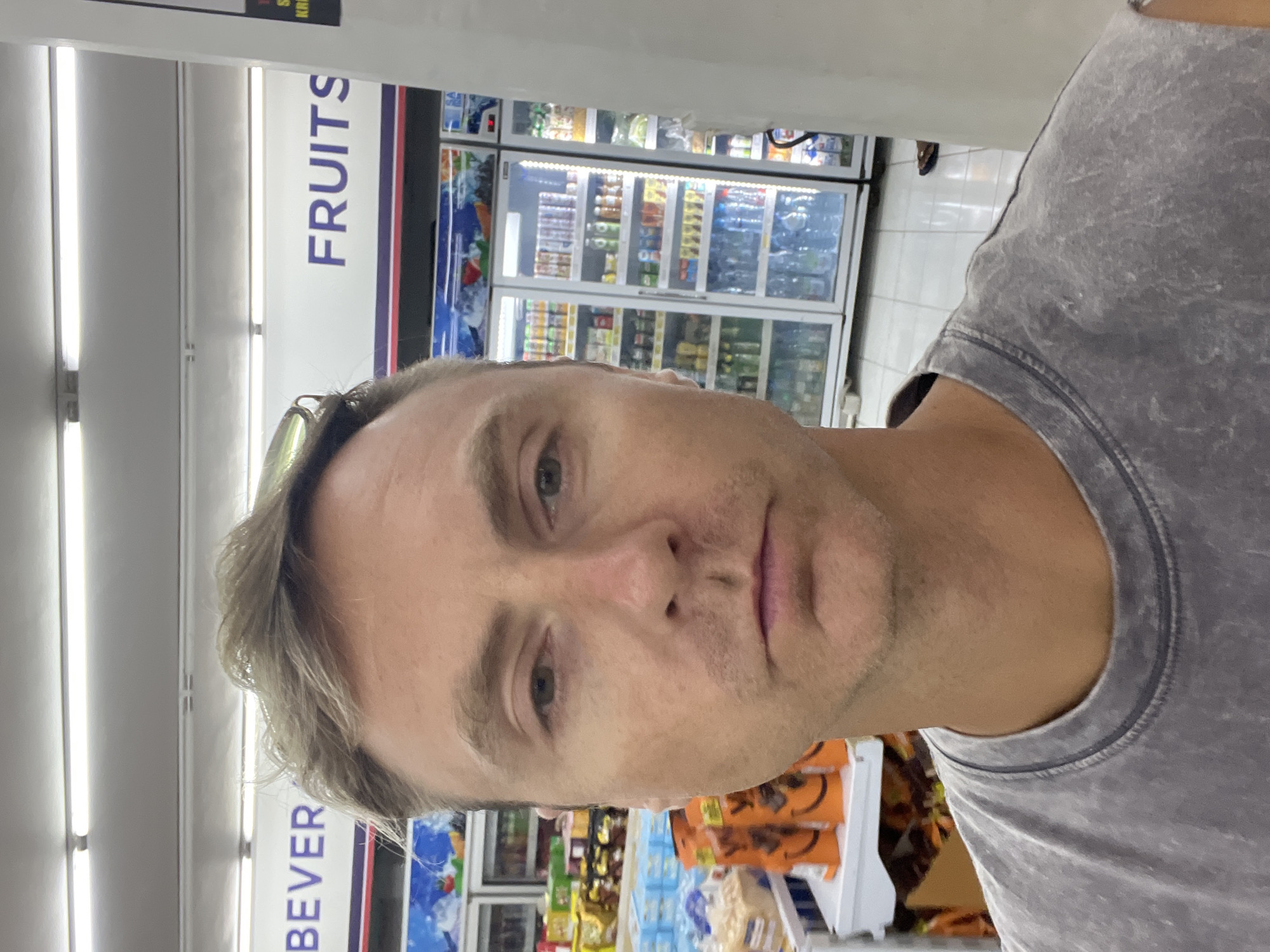 The image shows a man in a gray shirt, likely a selfie, inside a grocery store in Sukawati, Indonesia. The store has sections for "FRUITS" and "BEVERAGES" as indicated by signs on the walls. Refrigerated display cases filled with drinks are visible in the background. Aisles with various snack and food products can also be seen. The man appears to be standing near shelves containing products like candies and packaged foods. The store has typical fluorescent lighting.
