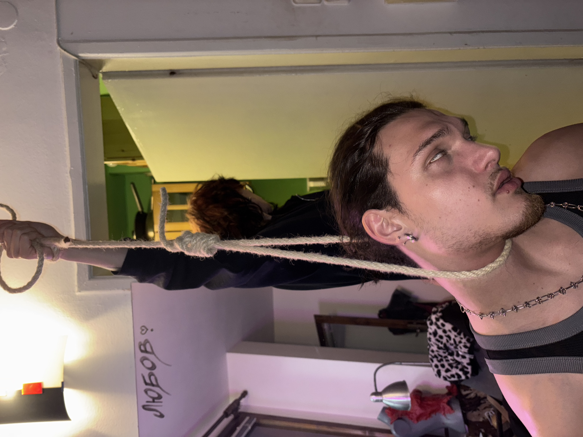 A close-up image showing a person with light skin, dark hair, and a beard, seemingly suspended by a rope tied around their neck. Their eyes are rolled back, and their mouth is slightly open. They are wearing a gray and black tank top and a silver chain necklace. A rope is looped around their neck and held by another person with auburn hair in the background, who is mostly obscured. The rope is also looped around the top of a doorway. The background includes a room with various items, including a desk lamp and a white board with the word "любов" written on it. The location is potentially in Sofia, Bulgaria.