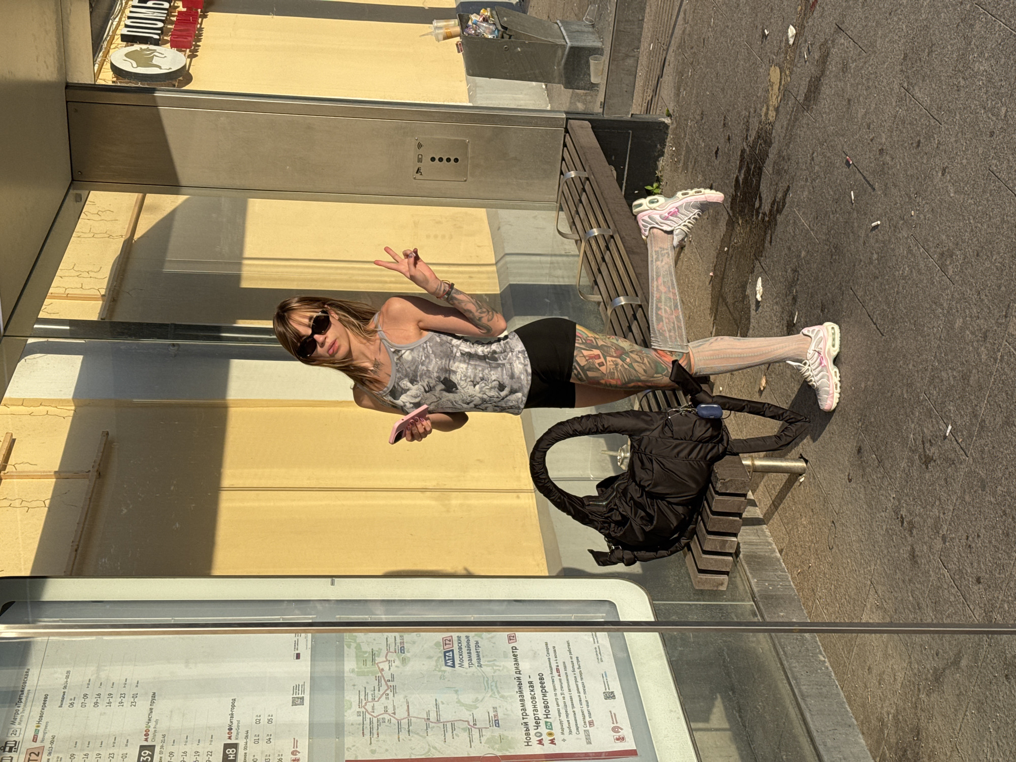 In Moscow, Russia, at a bus or tram stop, a young woman with extensive tattoos is posing for a photo. She is wearing sunglasses, a patterned tank top, black shorts, and white socks with sneakers. Her left leg is raised, and she is holding a pink smartphone in her right hand while making a peace sign with her left. A black duffel bag rests on a wooden bench beside her. The background features a yellow wall with a sign displaying a bull logo and Cyrillic text. The ground is paved with stone tiles, and there is visible litter, including cigarette butts and discarded cups. A public transport schedule is displayed on a panel to the left of the woman. The scene is illuminated by warm, late afternoon sunlight.