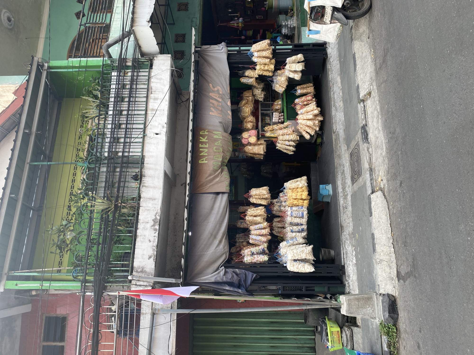 The image depicts a street-level view of a small shop in Kediri City, Indonesia, specializing in snacks. The shopfront is open, displaying an assortment of bagged and strung snacks, likely crackers or chips, hanging from the awning and within the shop. A banner above the entrance reads "ANEKA MAKAN KHAS," which translates to "Various Local Snacks." The shop is flanked by a green metal door on the left and a more colorful interior on the right, suggesting other businesses or living spaces. A red and white Indonesian flag is visible, attached to a pole on the left side of the shop. The street in front of the shop is paved, with a concrete sidewalk and curb. A portion of a parked motorcycle is visible on the right side of the frame. The buildings in the background show multiple stories with balconies and various colored facades, indicative of urban architecture in Indonesia. The overall scene is that of a typical commercial street in an Indonesian city.