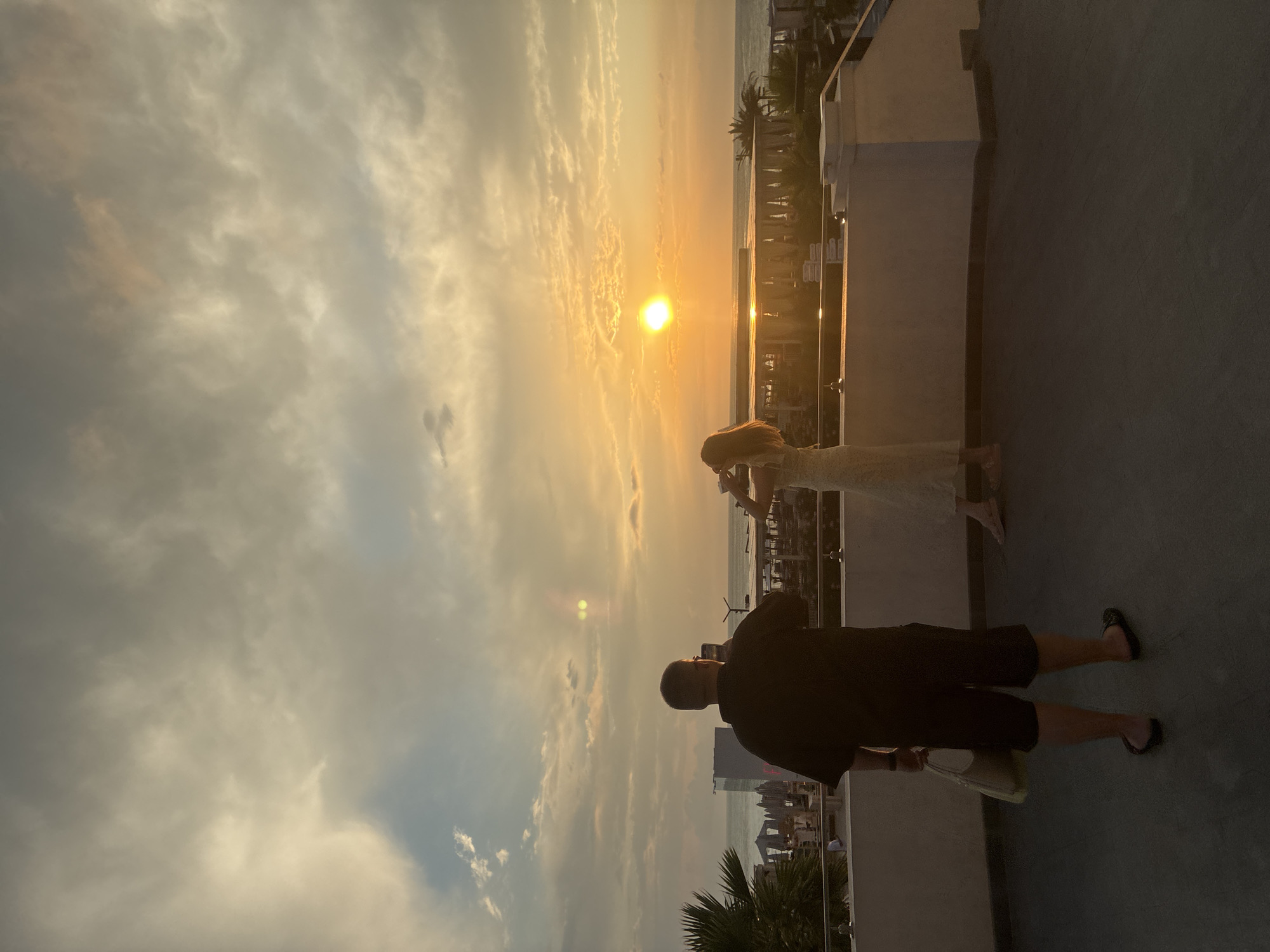 Two individuals are standing on a rooftop terrace overlooking the Black Sea at sunset. The man on the left is facing away from the camera, wearing a black t-shirt and dark shorts, holding a smartphone to take a photo. He is also carrying a light-colored tote bag. The woman on the right is also facing away, wearing a light-colored dress, and appears to be taking a photo or looking at the sunset through her phone. The sun is setting over the water, casting a warm glow across the sky and clouds. The foreground of the terrace features a dark tiled floor and a white railing. In the background, there are palm trees, lounge chairs, and buildings along the coastline, suggesting a resort or hotel setting in Sochi, Russia.