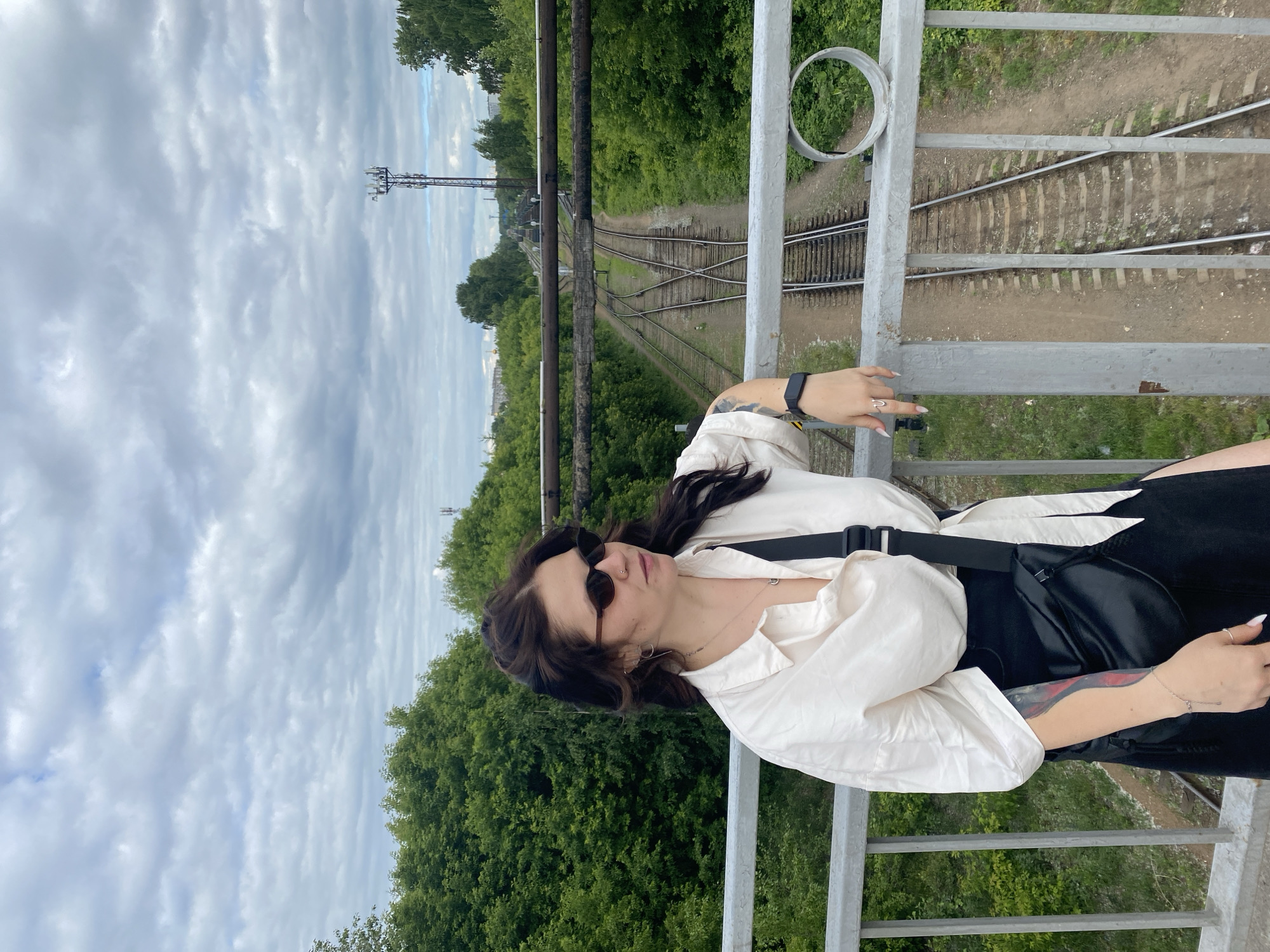 A young woman with dark hair and sunglasses stands on a bridge overlooking train tracks in Chaikovsky, Russia. She is wearing a white button-down shirt, a black skirt, and a black crossbody bag. The train tracks run through a lush green forest, with a cell tower visible in the distance. The sky is cloudy.