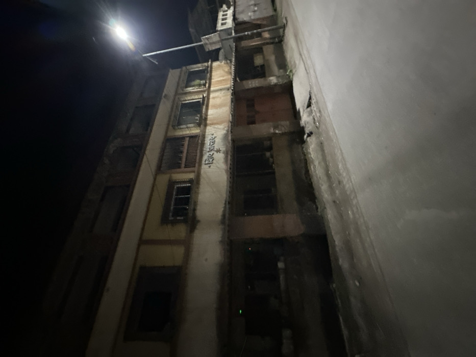 A dimly lit urban scene presents a narrow alleyway between a tall, deteriorating building and a plain wall. The building has multiple stories with some windows boarded up or missing. Visible writing appears on one section of the building's facade. A street lamp casts a bright glow at the top of the image, illuminating the top section of the buildings. The alley is shrouded in deep shadows, revealing structural elements of the building such as supports and bare walls.