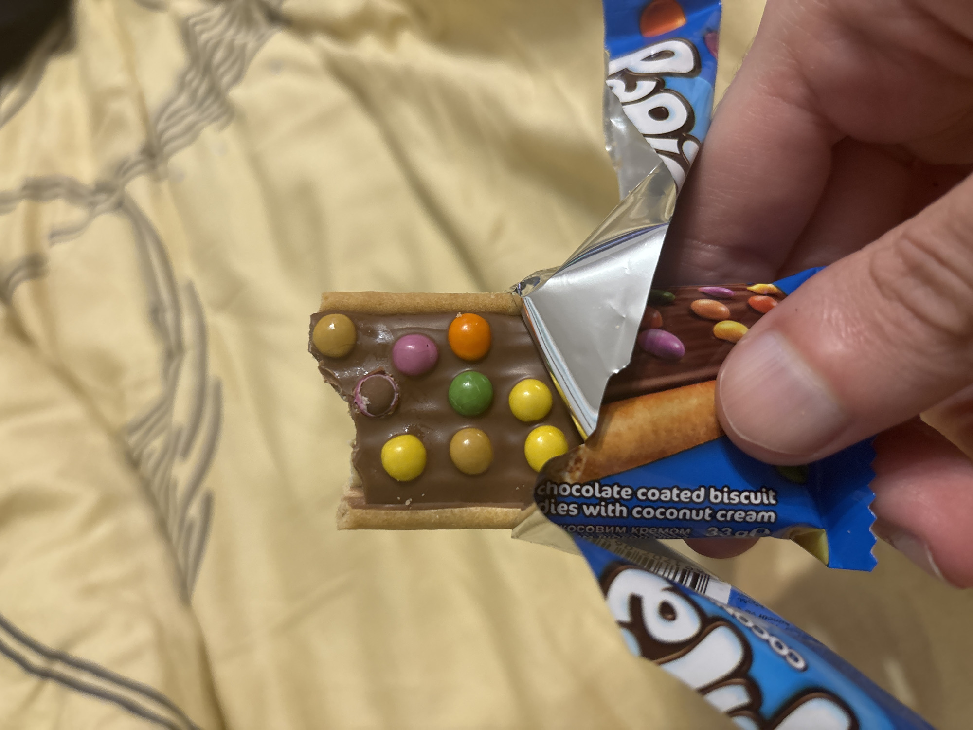A close-up, eye-level shot shows a person's hand holding a partially eaten chocolate-coated biscuit bar with colorful candy-coated chocolate pieces. The biscuit bar is rectangular and has a bite taken out of the top right corner. It is covered in milk chocolate and adorned with multiple round, brightly colored confectionery pieces in yellow, orange, green, pink, and brown. The biscuit bar is partially unwrapped from its silver foil packaging, revealing some blue packaging underneath with white lettering and graphics. The background consists of a light brown, textured fabric, possibly a blanket or bedding. The lighting is soft and even, with no harsh shadows.