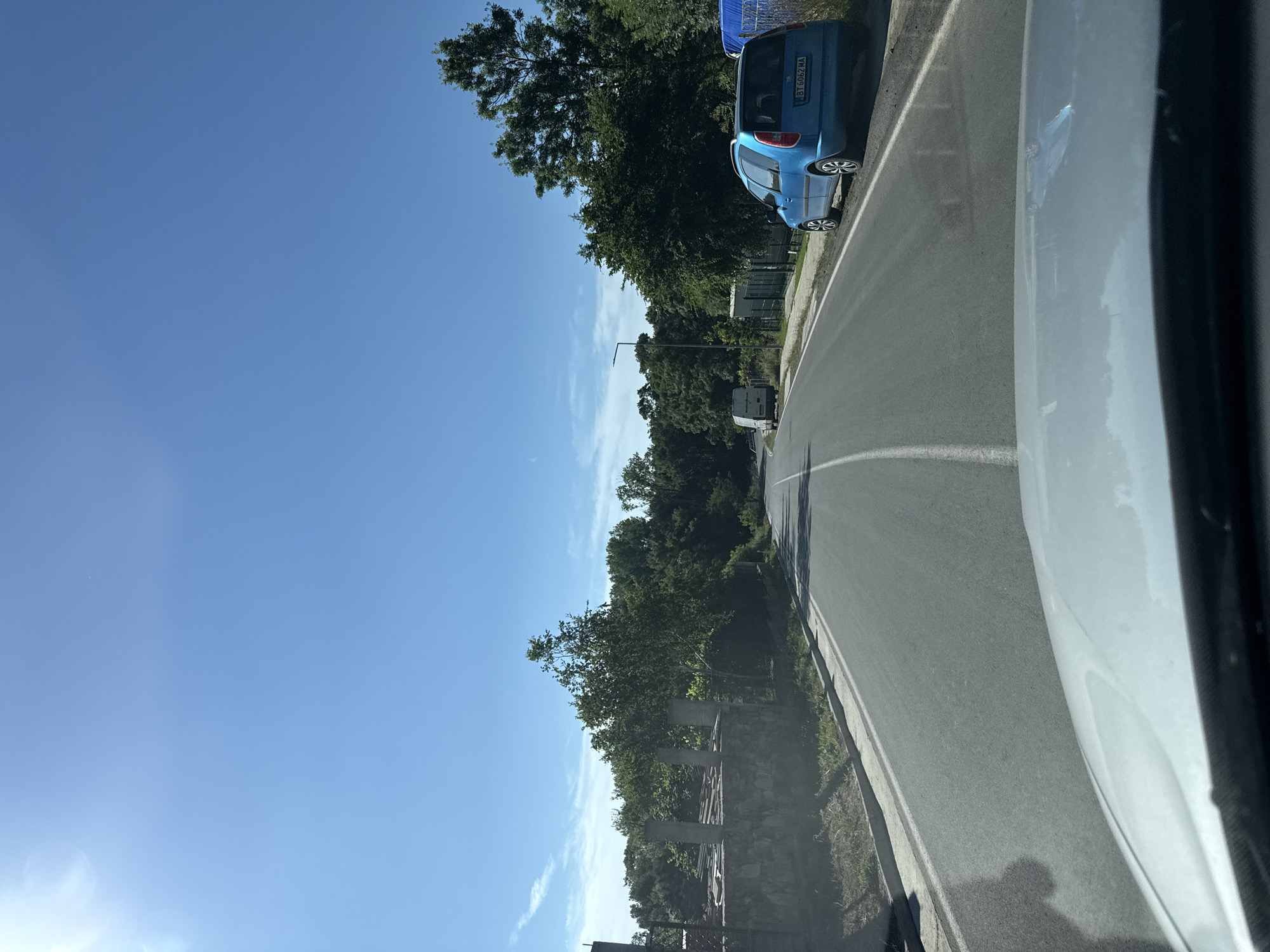 A clear blue sky with the sun creating lens flare is above a two-lane paved road. The road curves to the left, bordered by grass and a curb. On the right side of the road, a blue station wagon is parked facing away from the camera, its license plate is visible. Beyond the car, a fence separates the road from a dense line of green trees. On the left side of the road, a stone wall is topped with concrete pillars and what appears to be construction materials. Further down the road, a white van is visible in the distance. The scene is a roadside view on a sunny day, likely in a semi-rural or suburban area of Tsarevo, Bulgaria.