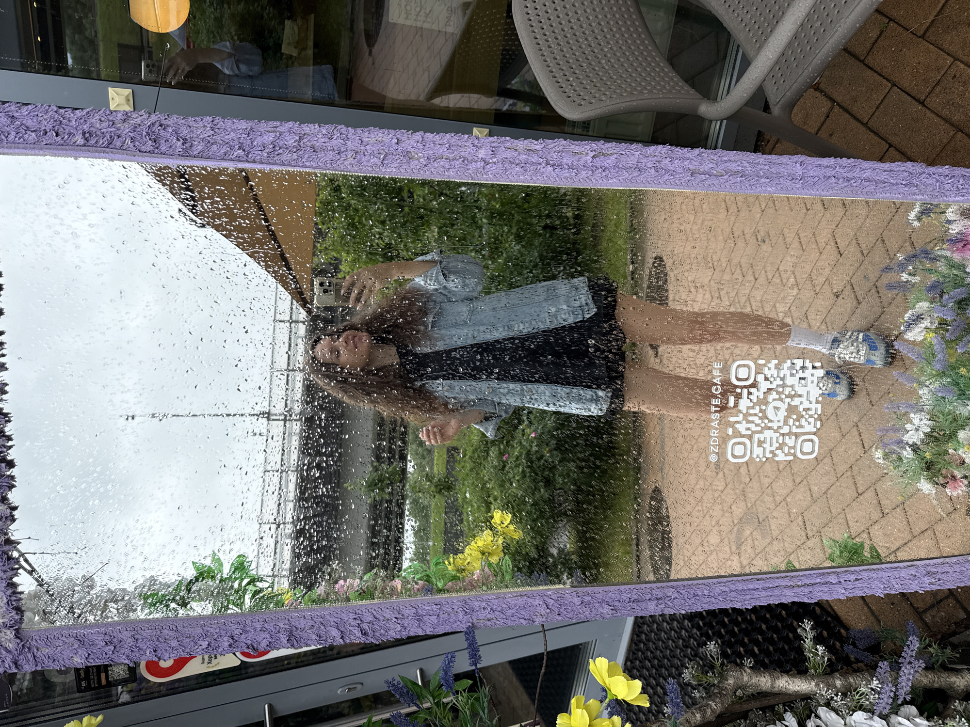 A young woman with curly brown hair is taking a selfie in a large, ornate mirror with a purple, textured frame. She is wearing a dark dress and a light blue denim jacket, and appears to be standing on a paved walkway. The mirror's surface is covered in raindrops, obscuring the background, which shows greenery and a cloudy sky. A QR code and the text "@ZDRASTE.CAFE" are visible on the mirror. The scene is likely outdoors, possibly near a cafe, in the city of Dolgoprudny, Russia, given the Cyrillic text on a sign attached to the mirror frame.