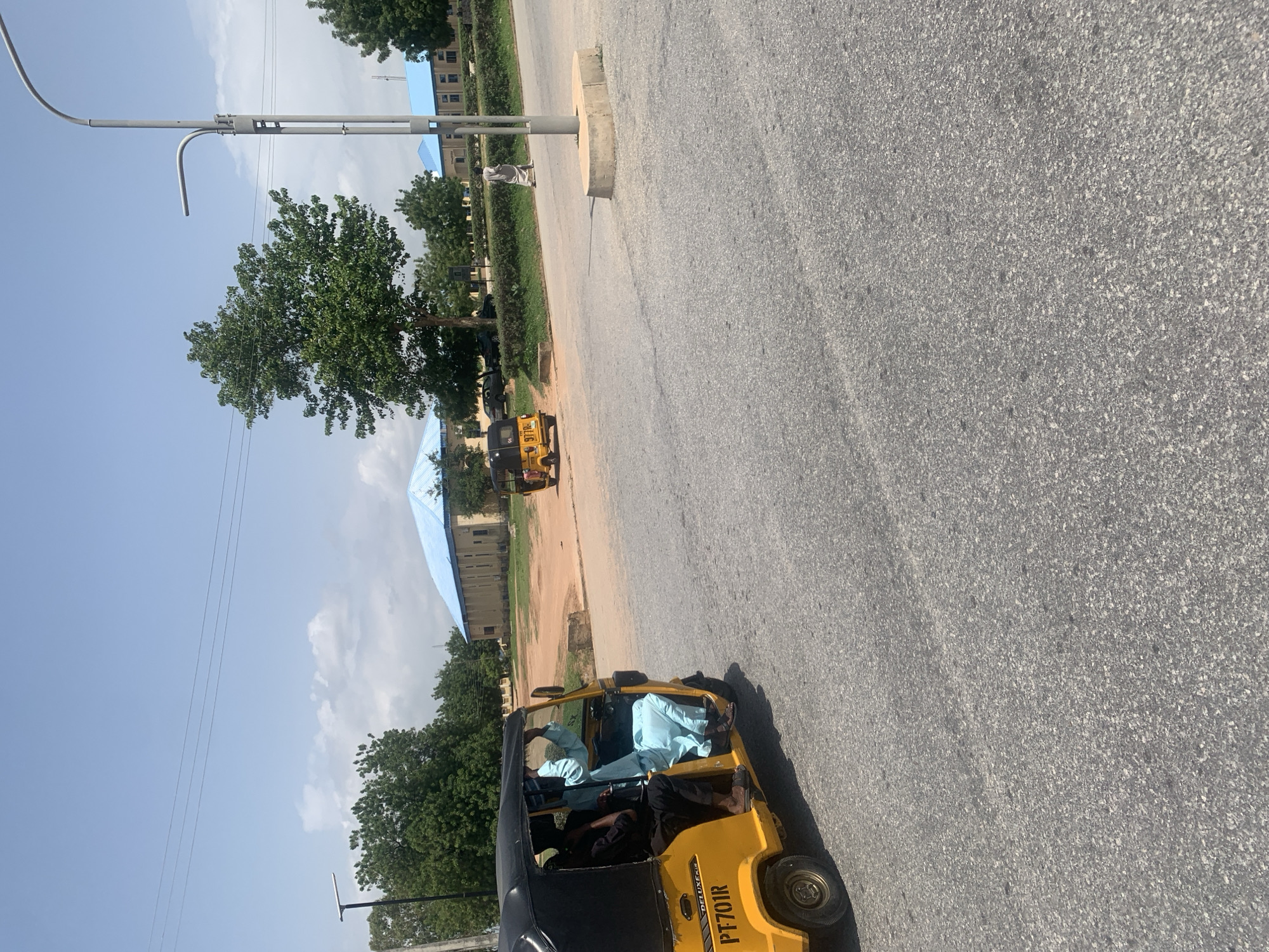 This is a photograph taken outdoors in Jalingo, Nigeria, likely during the daytime given the bright sunlight and shadows. The scene depicts a paved road with a yellow auto-rickshaw, also known as a "Keke Napep" or "Keke" in Nigeria, driving on it.
Inside the auto-rickshaw, at least two individuals are visible. The driver appears to be wearing dark clothing, while a passenger in the front is dressed in a light blue traditional garment. Another person is seated in the back, also wearing dark clothing. The auto-rickshaw has "PT701R" printed on its side.
In the background, there are buildings with light blue roofs, green trees, and some vegetation lining the side of the road. A lamppost is also visible. The sky is blue with some scattered clouds, suggesting fair weather. The overall impression is a snapshot of daily life on a road in a Nigerian urban or semi-urban environment. The photograph is oriented vertically, making the road appear to be running diagonally across the frame.