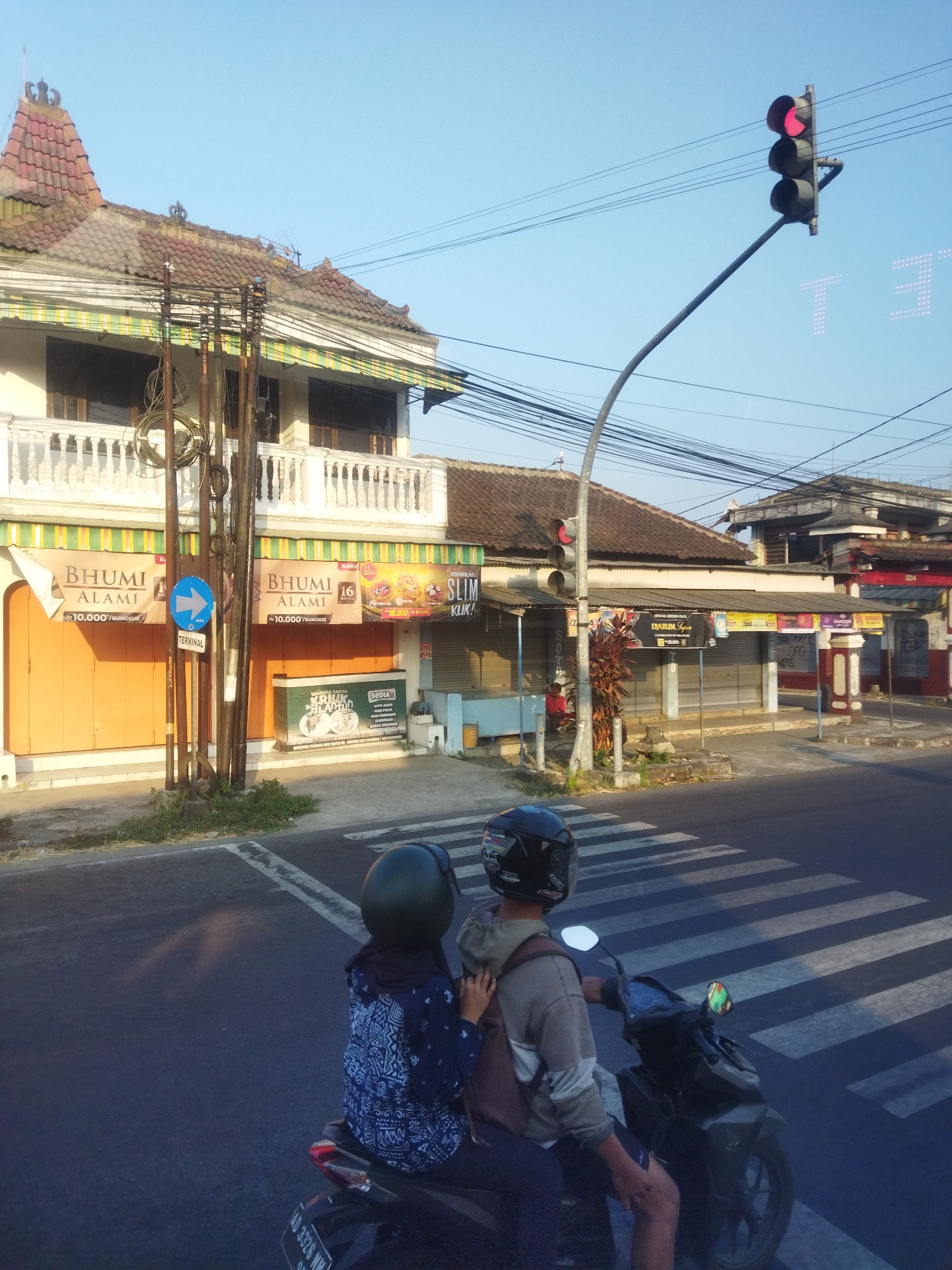 The image shows a street scene in Singodutan, Indonesia. A traffic light is red, indicating that vehicles must stop. Two people are on a motorcycle, stopped at a pedestrian crossing. The person in front is wearing a blue and white patterned shirt and a dark helmet, while the person behind is wearing a grey hooded sweatshirt and a black helmet. They are facing away from the camera. On the right side of the street, there are several buildings with closed storefronts. One building has a sign that says "BHUMI ALAMI." The overall environment appears to be a commercial area in the early morning or late afternoon, given the long shadows.