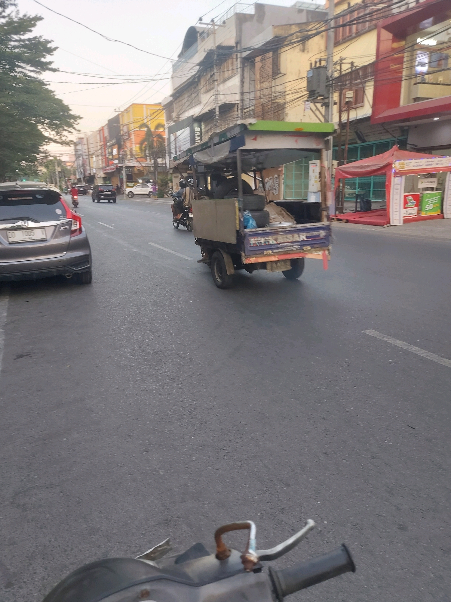 The image depicts a street scene in Makassar, Indonesia, likely during daylight hours with overcast skies. The perspective is from a vehicle moving on the road, focusing on the traffic and the buildings lining the street.
On the left side of the road, a dark-colored hatchback car is parked or moving slowly. Further down the road, a red scooter is visible. On the right side of the road, there is a small motorized truck with a covered bed, carrying tires and other items. Behind this truck, a red tent structure is set up on the sidewalk, possibly for a market stall or event. Several buildings of varying heights and architectural styles line the street, with some appearing to be commercial establishments. The road surface is asphalt, marked with white lane dividers. Electrical wires are strung overhead, a common sight in urban Indonesian environments. The general atmosphere is that of a busy, active urban street.