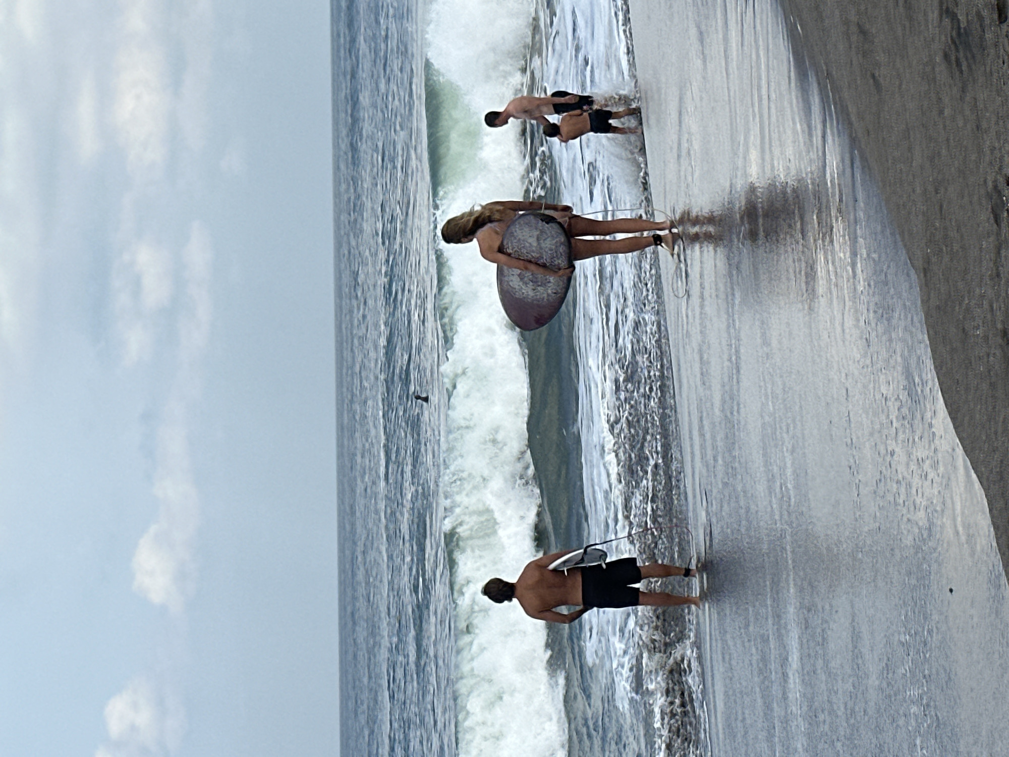In North Kuta, Indonesia, four individuals are at the edge of the ocean on a beach. The sky is overcast with scattered clouds. A man on the left, facing away from the camera, wears black shorts and holds a white surfboard. To the right of him, a woman also facing away, holds a multi-colored surfboard. Further to the right, a man and a boy stand near the water's edge, with the boy appearing to be playing in the surf. Large waves are breaking in the ocean, and a dark shape, possibly a surfer or a marine animal, is visible in the distance. The wet sand reflects the gray sky.