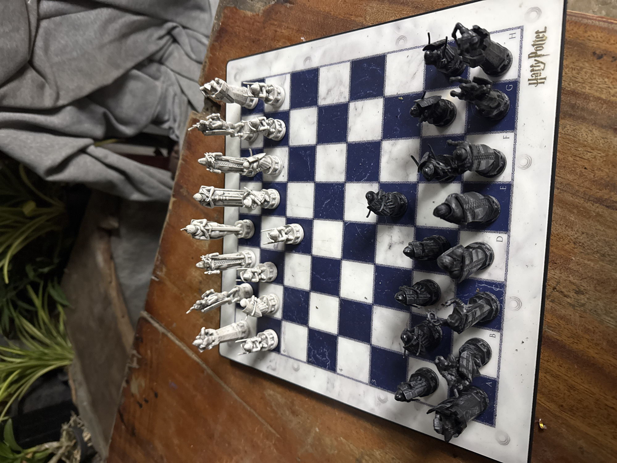 A Harry Potter chess set is arranged on a square chessboard with blue and white squares. The white pieces are on the left side of the board, while the black pieces are on the right. The chess board rests on a wooden surface. Part of a gray fabric and some greenery are visible in the background. The chess board has the words 'Harry Potter' written in the top right corner. The location of the image is not visible, and information about the city of Moo 4, Thailand, is not available.