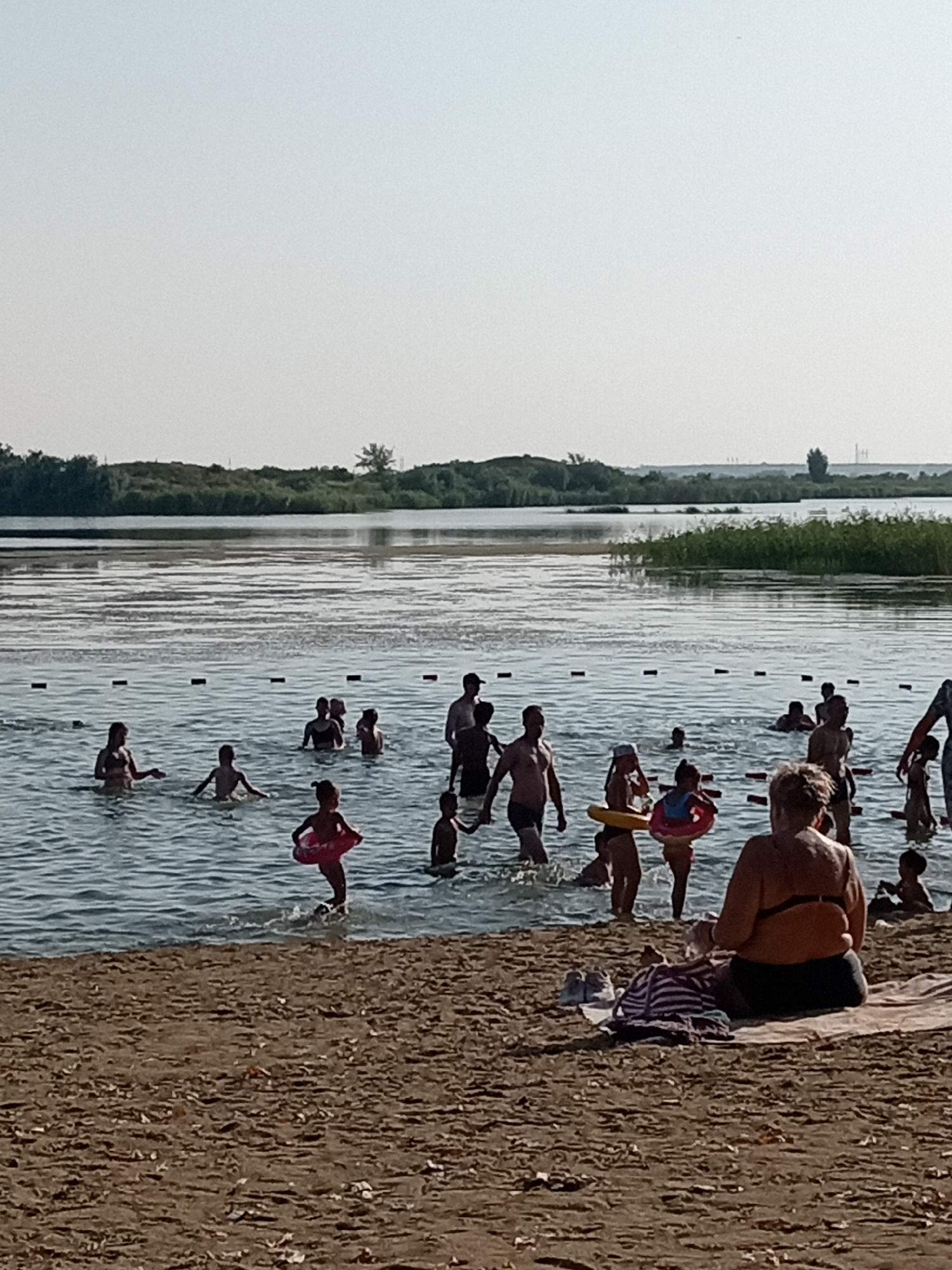 The image depicts a summer day at a beach or riverbank, likely in Rudny, Kazakhstan, with numerous people enjoying the water. In the foreground, a sandy beach stretches across the bottom of the frame. Several people are seated on the sand, with one individual in the lower right corner wearing a dark swimsuit and black bra. Beach towels and bags are visible on the sand.
The majority of the activity takes place in the water. A group of people, including children and adults, are swimming and playing in the water. Some are wading, while others are further out. Several children are visible with inflatable swim rings. In the middle ground, a marked boundary line with buoys separates shallow from deeper water.
The background shows a body of water, likely a river or lake, with a treeline and vegetation on the opposite bank. The sky is clear and bright, indicating sunny weather. The overall scene is one of recreational activity in an outdoor, waterside environment.
