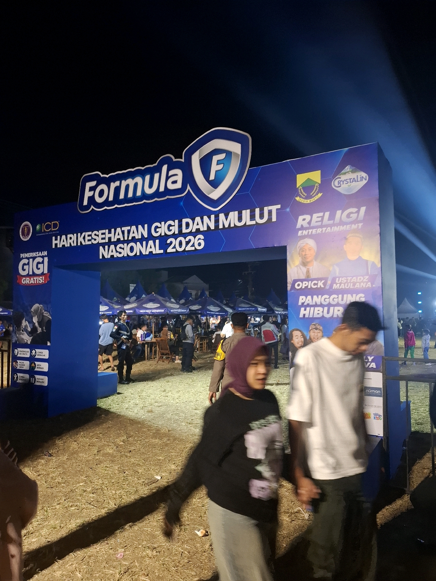 The image shows a nighttime outdoor event in Bojong, Indonesia, with a large blue archway entrance as the central focus. The archway is decorated with text and logos. Prominently displayed on the arch is "Formula F" and "HARI KESEHATAN GIGI DAN MULUT NASIONAL 2026" (National Oral and Dental Health Day 2026). Below this, a sign on the right side of the arch displays "RELIGI ENTERTAINMENT" with images of two men, identified as "OPICK" and "USTADZ MAULANA," and further text reading "PANGGUNG HIBUR" (Entertainment Stage). On the left side of the arch, a banner announces "PEMERIKSAAN GIGI GRATIS!" (Free Dental Check-up!).
Several people are visible in and around the archway. Two individuals are walking towards the camera in the foreground, a woman wearing a dark top and grey pants, and a man in a white t-shirt and dark pants. Behind them, a person in a security uniform stands near the entrance. Further back, tents are set up, suggesting a fair or festival. The ground is covered in dry grass and some scattered litter. The lighting appears to be artificial, with spotlights creating beams in the background. The overall impression is of a lively community event.
