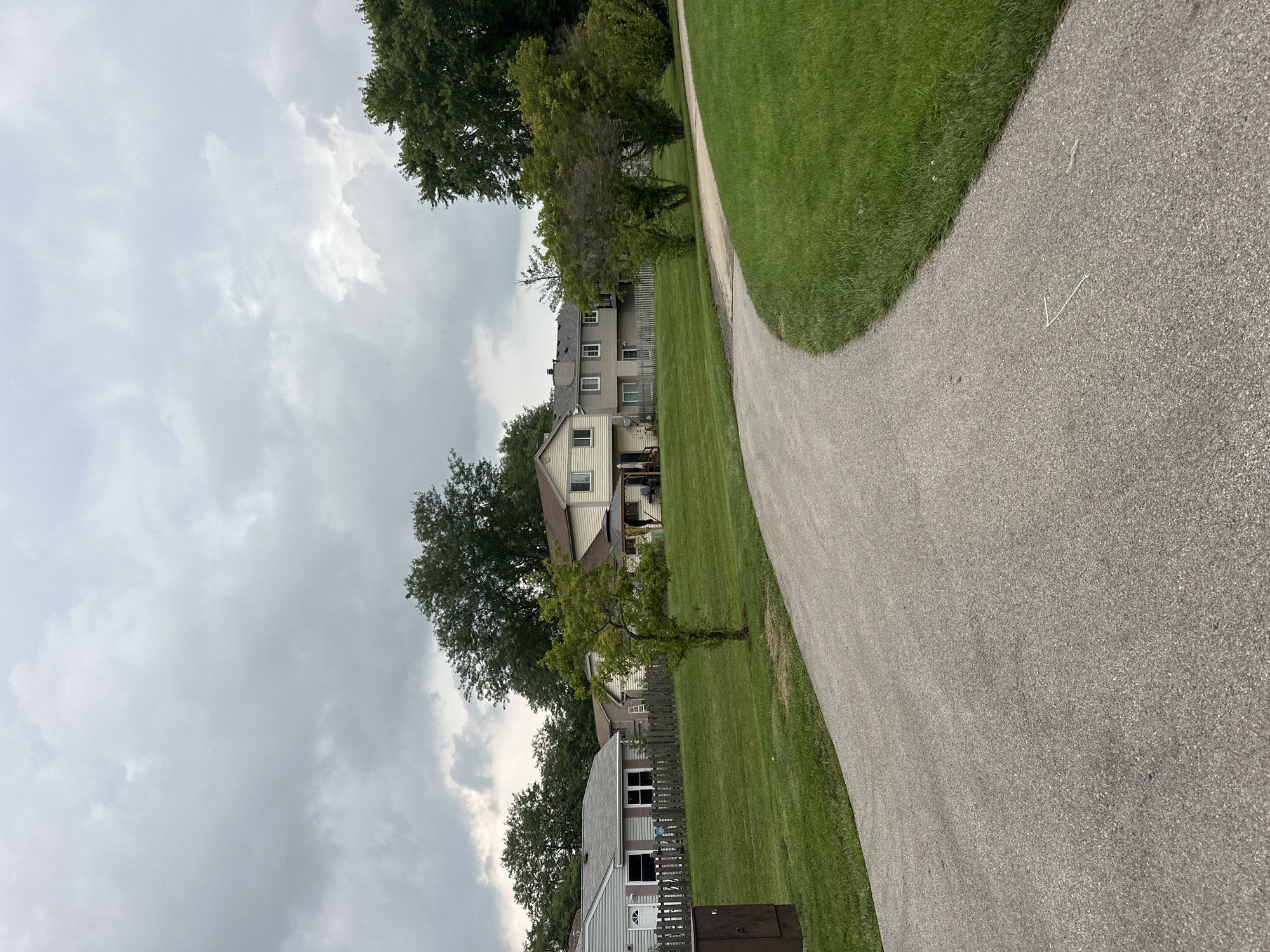 The image depicts a paved pathway curving through a grassy area in Streamwood, United States of America. Several residential buildings, possibly townhouses, are visible in the background, separated by a wooden fence. Tall trees surround the houses, and the sky is overcast with heavy gray clouds. A small, sparsely leafed tree stands in the middle of the lawn. A brown rectangular utility box is situated on the left side of the pathway. No people are visible in the scene. The overall impression is a quiet, suburban environment under a cloudy sky.