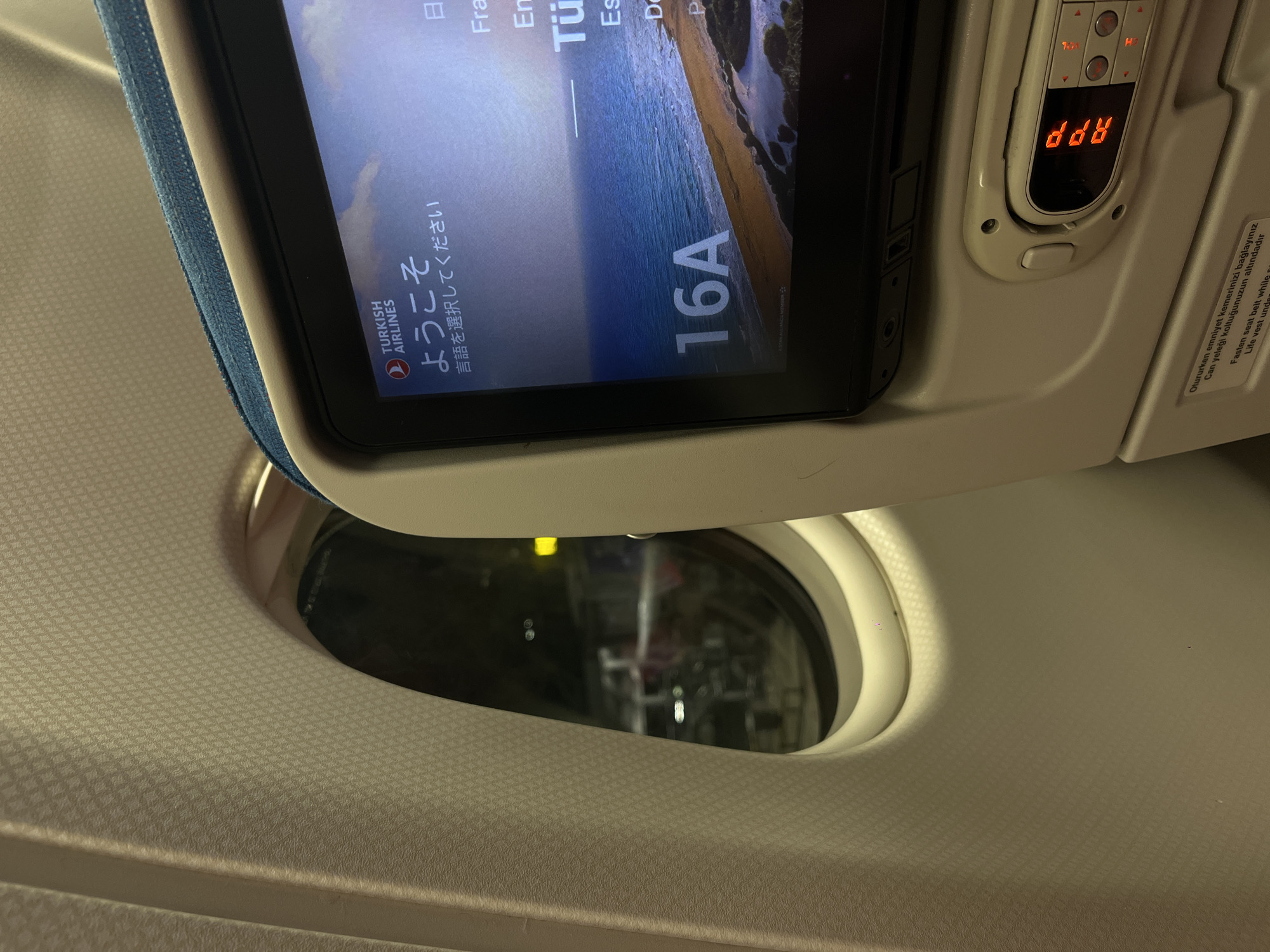 The image depicts the interior of an airplane cabin, specifically focusing on a passenger's seat. A window is visible on the left, showing a dark, blurred exterior that suggests nighttime or a tunnel. To the right of the window, a personal in-flight entertainment screen is displayed, showing the Turkish Airlines logo and "Welcome" in Japanese. The screen also displays the seat number "16A" and a scenic image of a coastline. Below the screen, the seat's control panel is visible, featuring buttons and a small digital display. The airplane's interior surfaces, including the seat upholstery and window frame, are visible. There are no people clearly visible in the image. The location in the city of Nong Prue Subdistrict, Thailand is not discernible from the visual information provided.