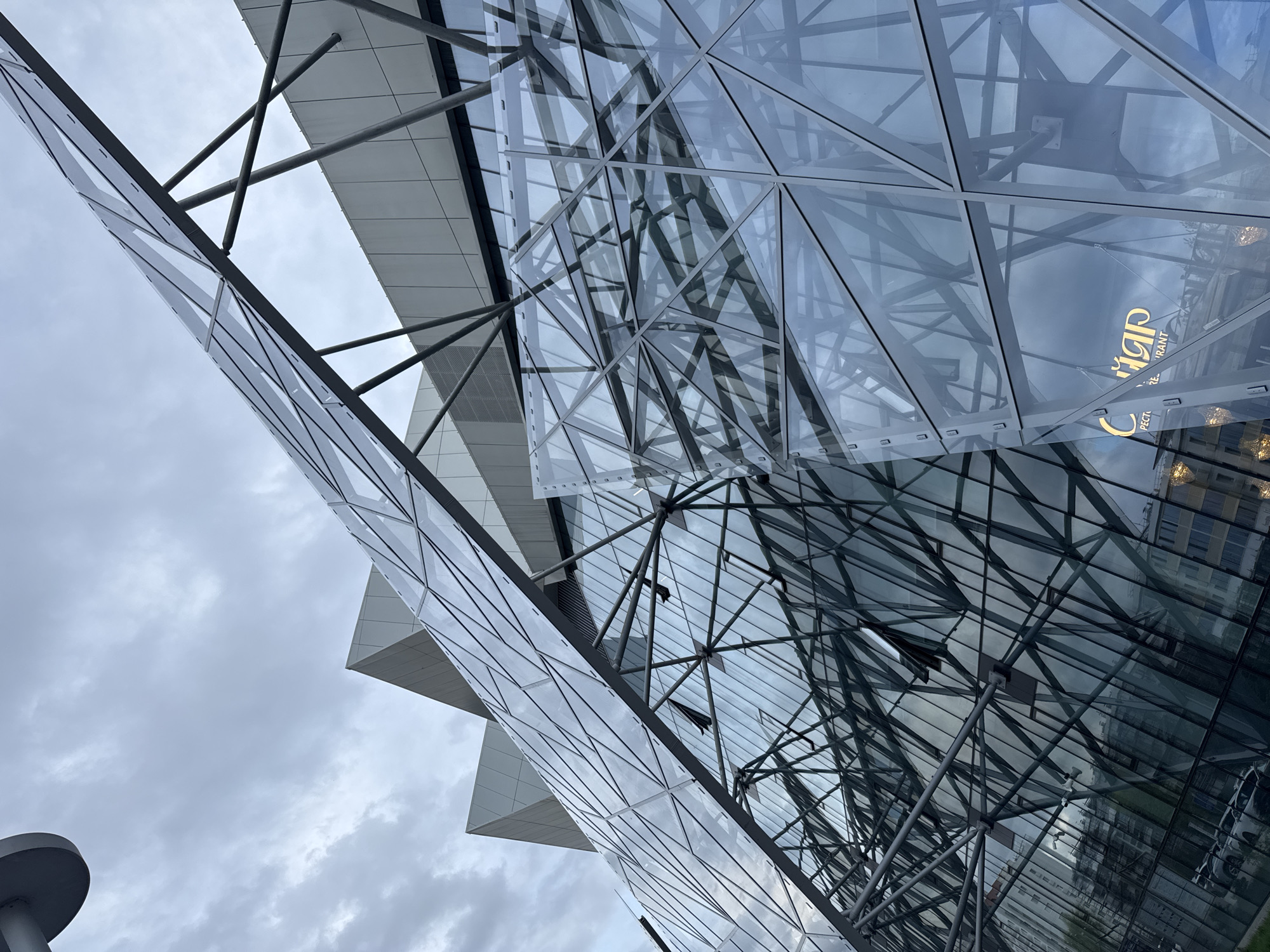 The image shows a low-angle view of a modern building with a complex glass and metal structure, likely located in Kazan, Russia, based on the reflection of the 'Cigar Restaurant' logo in the glass. The building appears to have a geometric design with triangular and polygonal glass panels supported by a network of metal beams and struts. The sky is overcast, with cloudy conditions reflecting on the building's glass surface. In the bottom left corner of the image, there is a partial view of a grey, possibly concrete object, potentially a street lamp or bollard. The perspective is tilted upwards, emphasizing the height and complexity of the architectural design. The reflection shows the surrounding buildings.