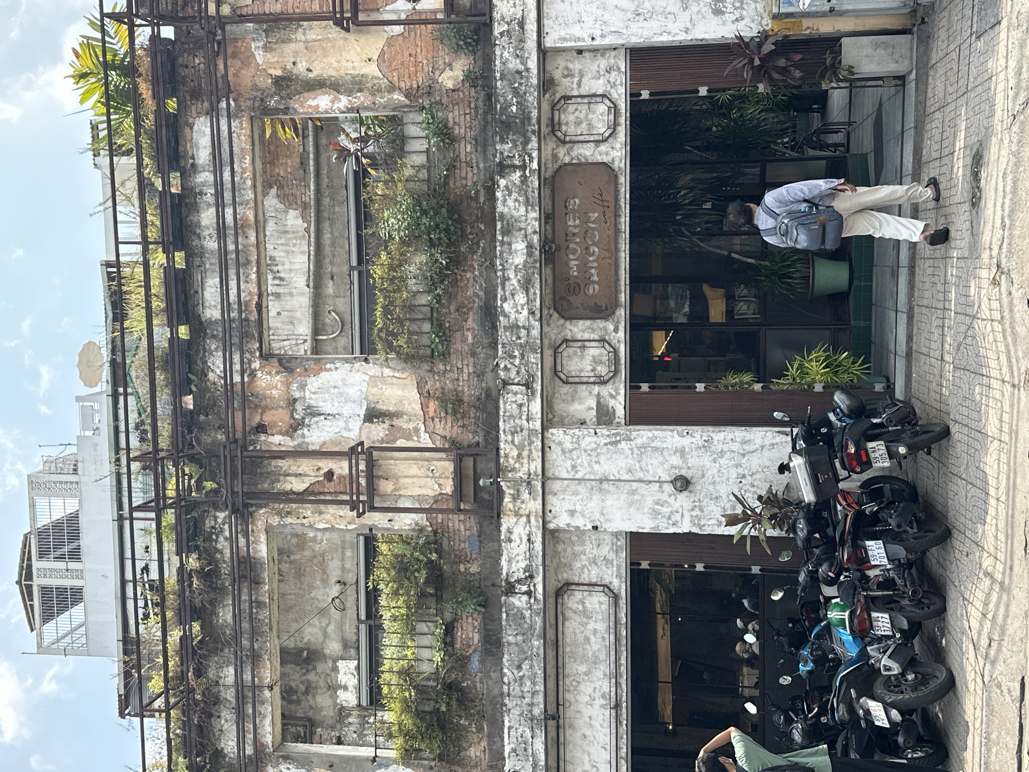 A vertical, full outdoor shot depicts the exterior of S'mores Saigon caffe in Thủ Đức, Vietnam. To the left, a derelict building shows decaying brick and concrete construction with open window spaces overtaken by vegetation and metal scaffolding. To the right is the functioning business front, featuring a sign bearing the cafe's name and closed, dark-tinted windows. A man with a backpack stands on the sidewalk near the entrance. Parked in front of the cafe are several scooters, their license plates bearing Vietnamese characters and numerals. The environment suggests an intersection of dilapidated infrastructure and contemporary urban commerce.