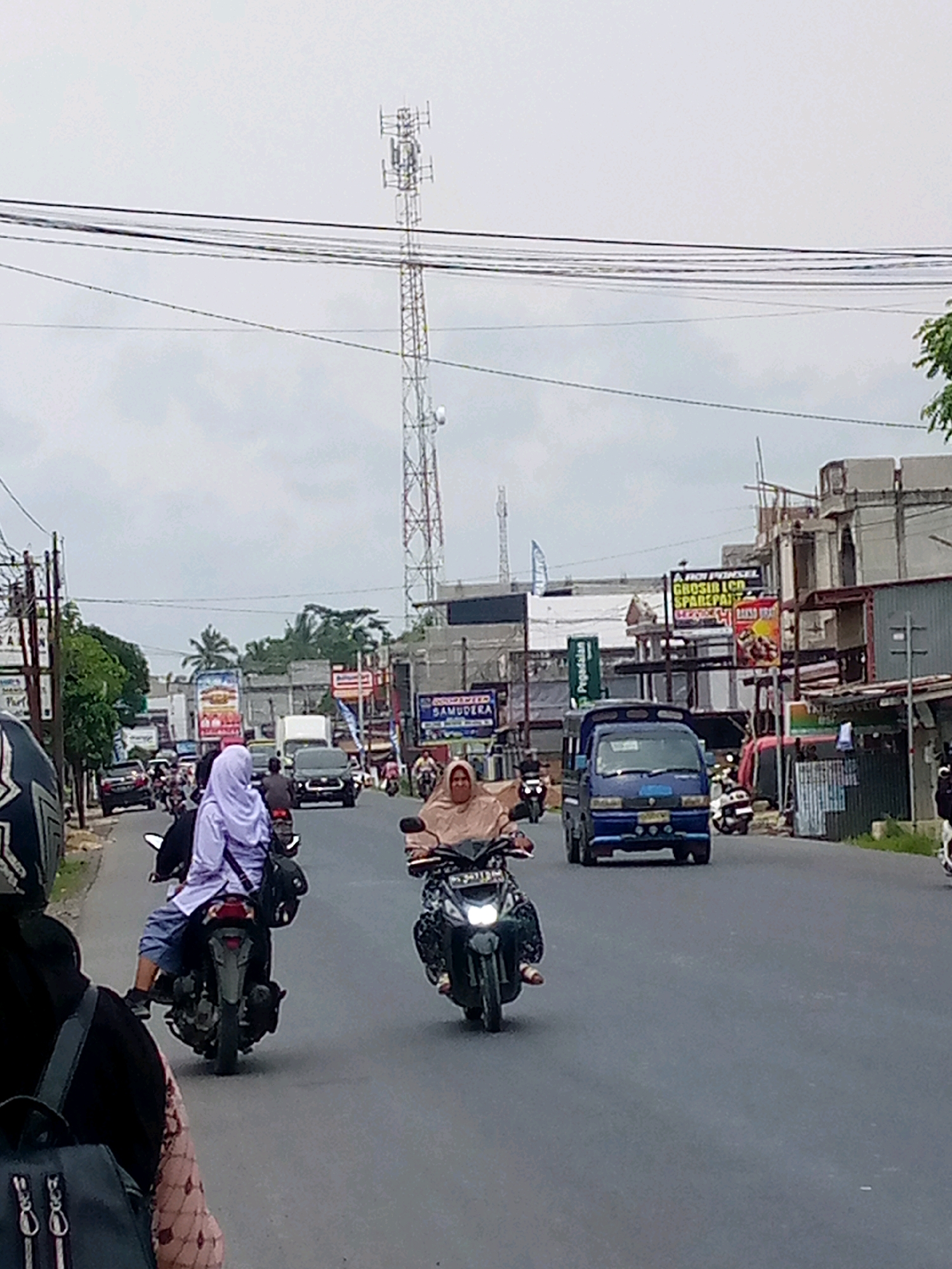 The image depicts a street scene in Keudebayu, Indonesia, under an overcast sky. In the foreground, a person wearing a black helmet and backpack is partially visible, facing away from the camera. They are on a motorcycle, indicated by the handlebars and part of the seat. To their right, another motorcyclist wearing a light purple hijab and blue pants is riding a black motorcycle. Further down the road, a person wearing a light brown hijab is riding a motorcycle, facing the camera. In the background, a blue utility truck is driving in the same direction as the motorcycles. Several other vehicles, including cars and motorcycles, are present on the road, indicating moderate traffic. Buildings line both sides of the street, with a prominent cell tower visible in the distance. Some buildings display advertisements and signs. The road surface is asphalt and appears wet in places, suggesting recent rain. The overall environment appears to be a commercial or mixed-use area within the town.