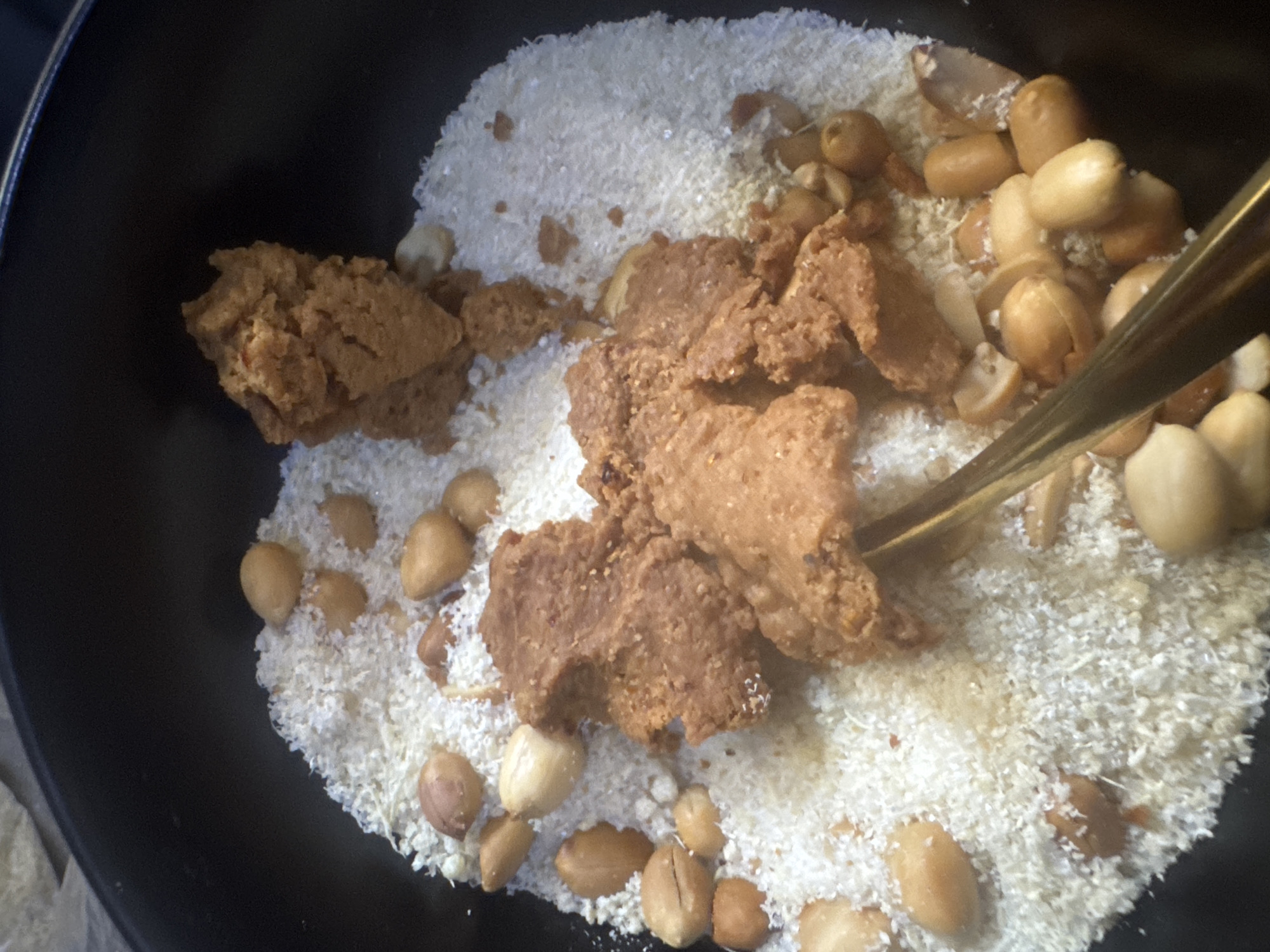 The image shows a close-up of ingredients for a Nigerian dish, possibly a groundnut soup or stew, being prepared in a dark pan. The main subjects are a pile of white, shredded coconut and several whole, shelled peanuts. Scattered amongst the coconut and peanuts are chunks of what appears to be groundnut paste, a common ingredient in West African cuisine. A golden utensil, possibly a spoon or a skewer, is visible in the upper right portion of the frame, interacting with the ingredients, suggesting the start of a mixing or cooking process. The lighting is bright, highlighting the textures of the ingredients. The overall setting is domestic, likely a kitchen, with the dark pan providing a stark contrast to the light-colored ingredients. There is no visible text in the image. The context of Abuja, Nigeria, suggests this could be a home-cooked meal preparation.
