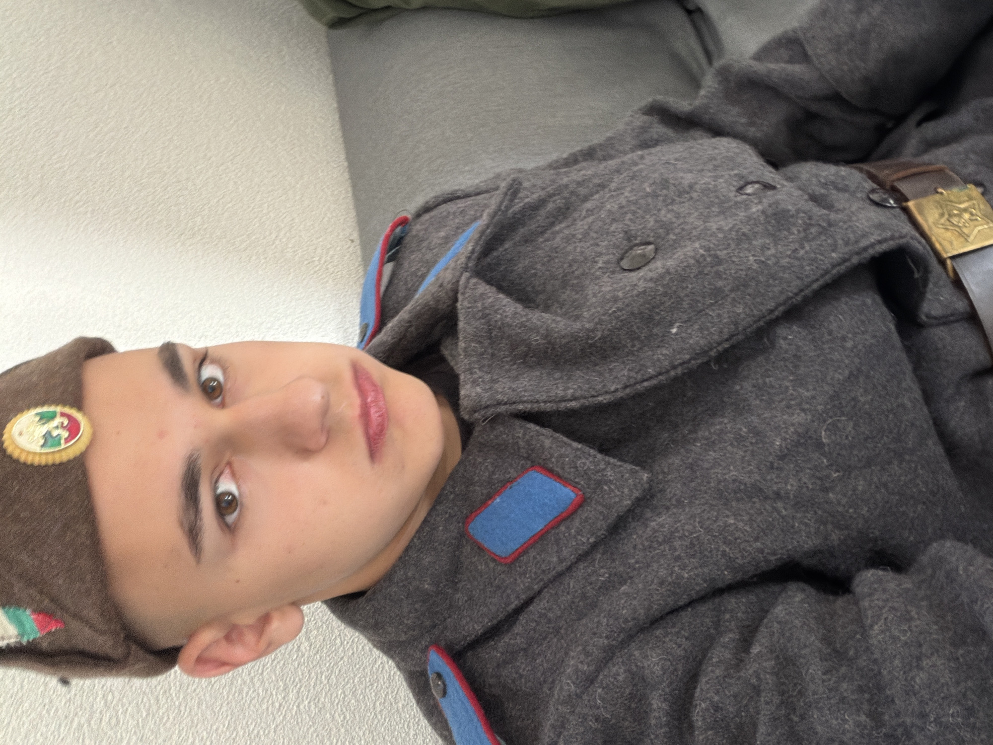 A young man is lying down, wearing what appears to be a vintage military uniform with a brown belt and a gold buckle. The uniform is gray with blue and red trim. He is also wearing a brown cap with a badge on it. The background is a plain wall and a gray surface. The location is unspecified in the image and there is no reference to Shumen, Bulgaria.