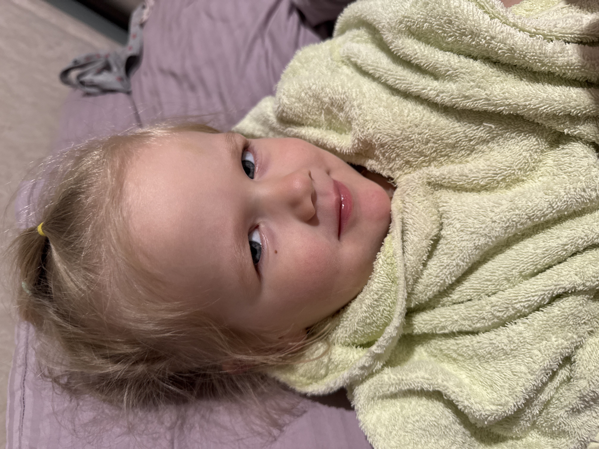A young blonde-haired child, appearing to be a toddler, is depicted lying down indoors, likely in a home setting. The child is wrapped in or covered by a soft, light green, terry cloth-like towel or blanket, which is draped around their body and partially covers their chin and neck.
The child's face is central to the image, with their head slightly tilted. They have bright blue or grey eyes, looking upwards and slightly to the right of the viewer. A gentle, faint smile plays on their lips, which appear soft and slightly glossy. There's a small, dark mole or freckle visible near the outer corner of their right eye. Their blonde hair is pulled back into a small ponytail on the right side of their head, secured with a yellow elastic band.
The background consists of blurred purple or lavender fabric, likely bedding or another blanket, suggesting a relaxed indoor environment such as a bedroom. Another very blurred, light-colored object, possibly clothing, is vaguely visible in the upper background.
The lighting is soft and ambient, characteristic of indoor conditions, creating a warm and peaceful atmosphere. The child's expression conveys contentment and serenity. There is no visible text in the media file.