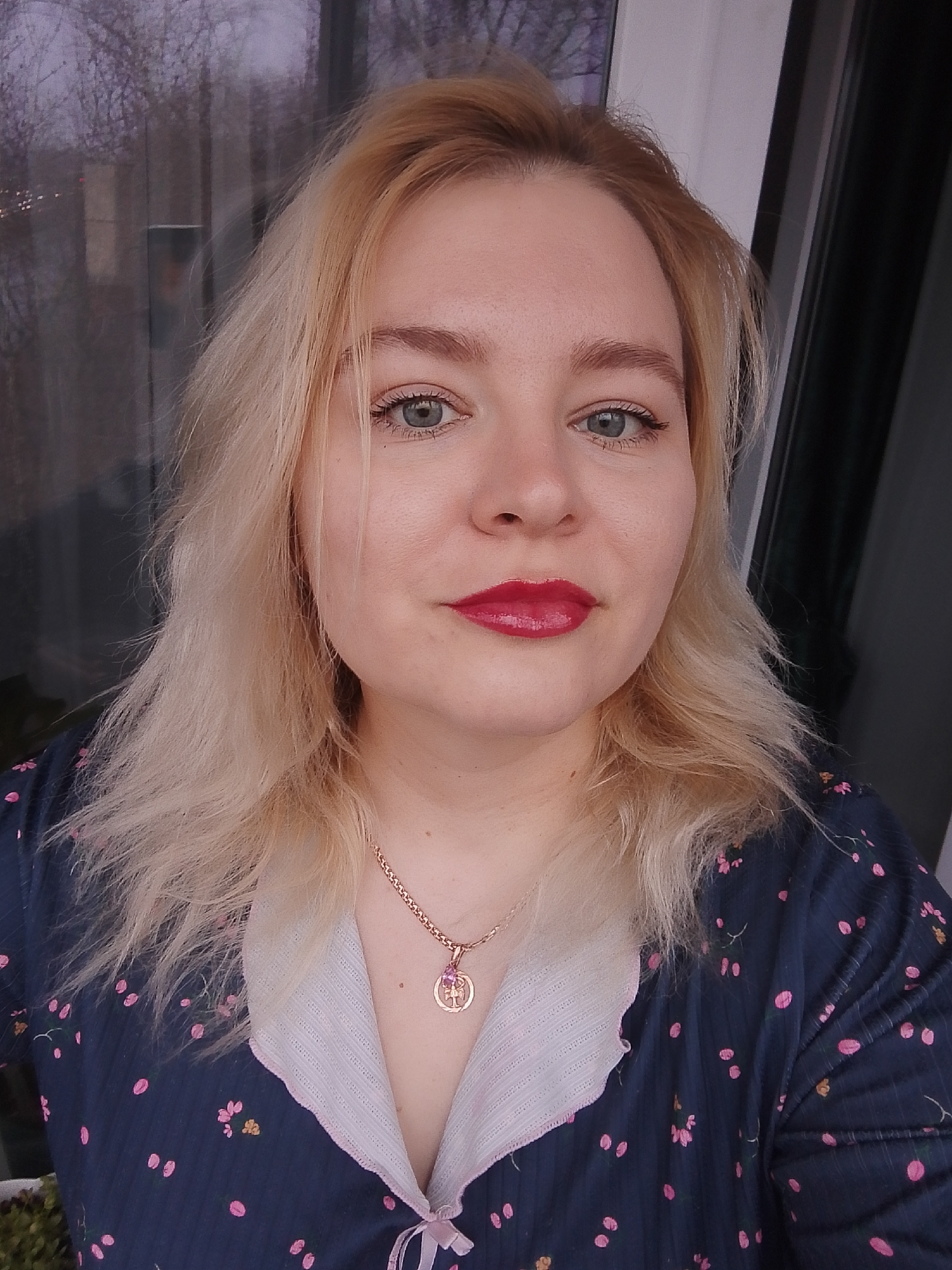 A Caucasian woman with blond, shoulder-length hair and blue-gray eyes is centered in the frame. She wears bright red lipstick and is gazing directly at the camera. Around her neck is a gold necklace with a circular pendant. She is wearing a dark blue pajama top with a white collar and small pink flower patterns, tied at the front with a light pink bow. Behind her, a window reflects an overcast cityscape. It could be determined to be a place in Izhevsk, Russia through contextual information. The overall impression is of a casual indoor setting, likely a personal residence.