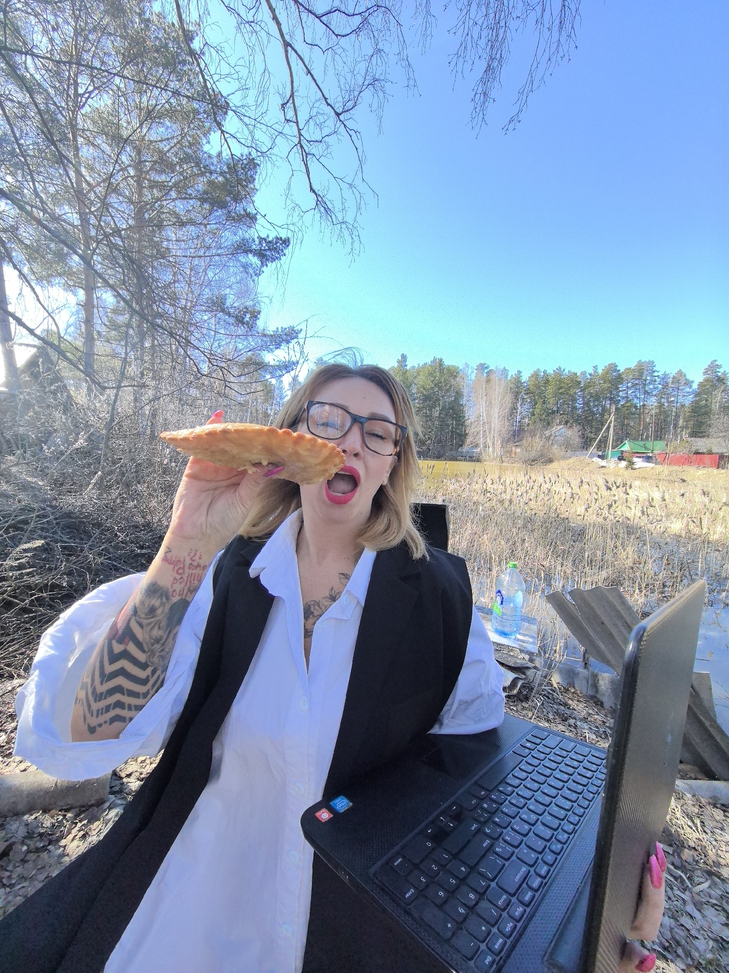 The image shows a woman outdoors on what appears to be a sunny day. She is wearing glasses, a white shirt, and a black vest, and has tattoos on her left arm and chest. She is holding a pastry and about to take a bite. There is a laptop in front of her, and a bottle of water is visible in the background. The environment includes trees, tall grass, and what appears to be a body of water. In the background, there are some buildings, including one with a red roof. Based on the context and information available, the location is identified as being in or near Noviy, Russia.