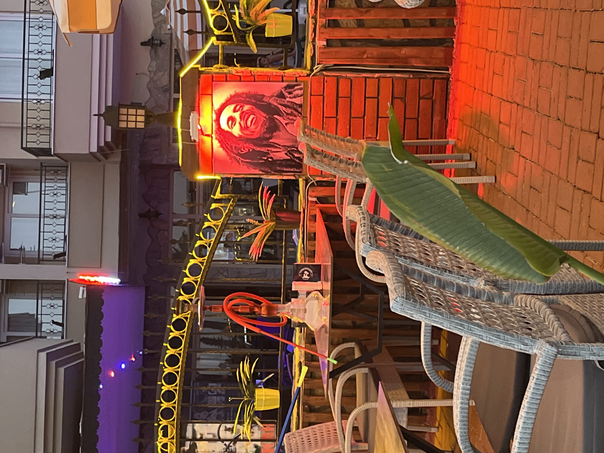 The image captures a vibrant outdoor scene in Alanya, Turkey. A large poster of Bob Marley, illuminated with red light, is prominently displayed against a brick wall. A decorative hookah with a red hose stands nearby. The foreground features wicker-style outdoor seating with a large green leaf partially obscuring the view. In the background are buildings with windows, a decorative iron fence adorned with string lights, and various plants in yellow pots. The overall atmosphere is lively and colorful, suggesting a restaurant or bar in a tourist area.