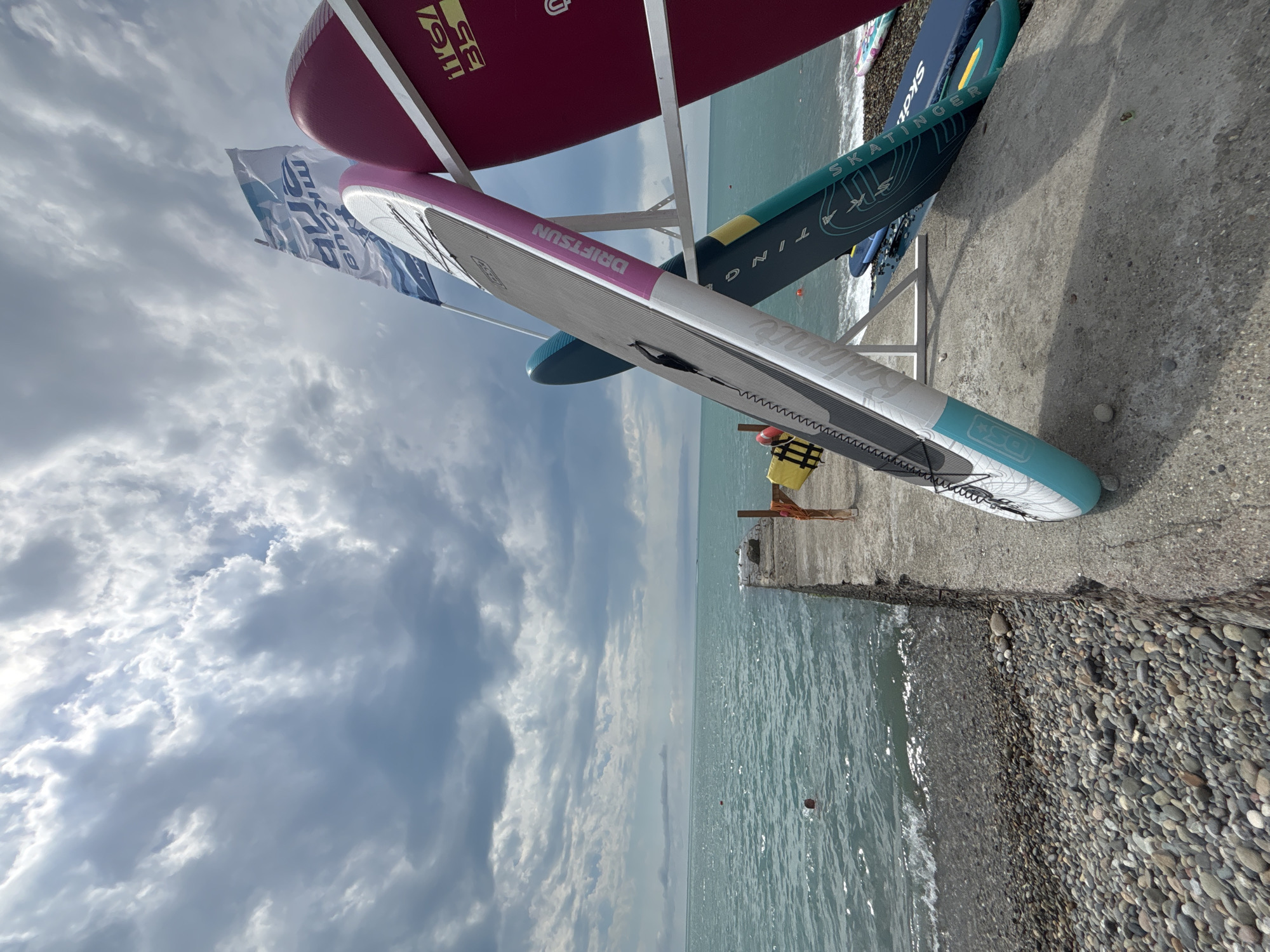 This is a low-angle, eye-level shot of a beach in Sochi, Russia. In the foreground, several stand-up paddleboards are propped up on a rack. One board is white with pink and turquoise accents, and another is dark red. Behind the paddleboards, the turquoise water of the Black Sea stretches to the horizon, meeting a cloudy sky. A concrete pier juts out into the water on the right side of the frame, with a person swimming in the distance. The beach is covered in grey and tan pebbles. A white flag with blue lettering is visible in the upper right portion of the image.