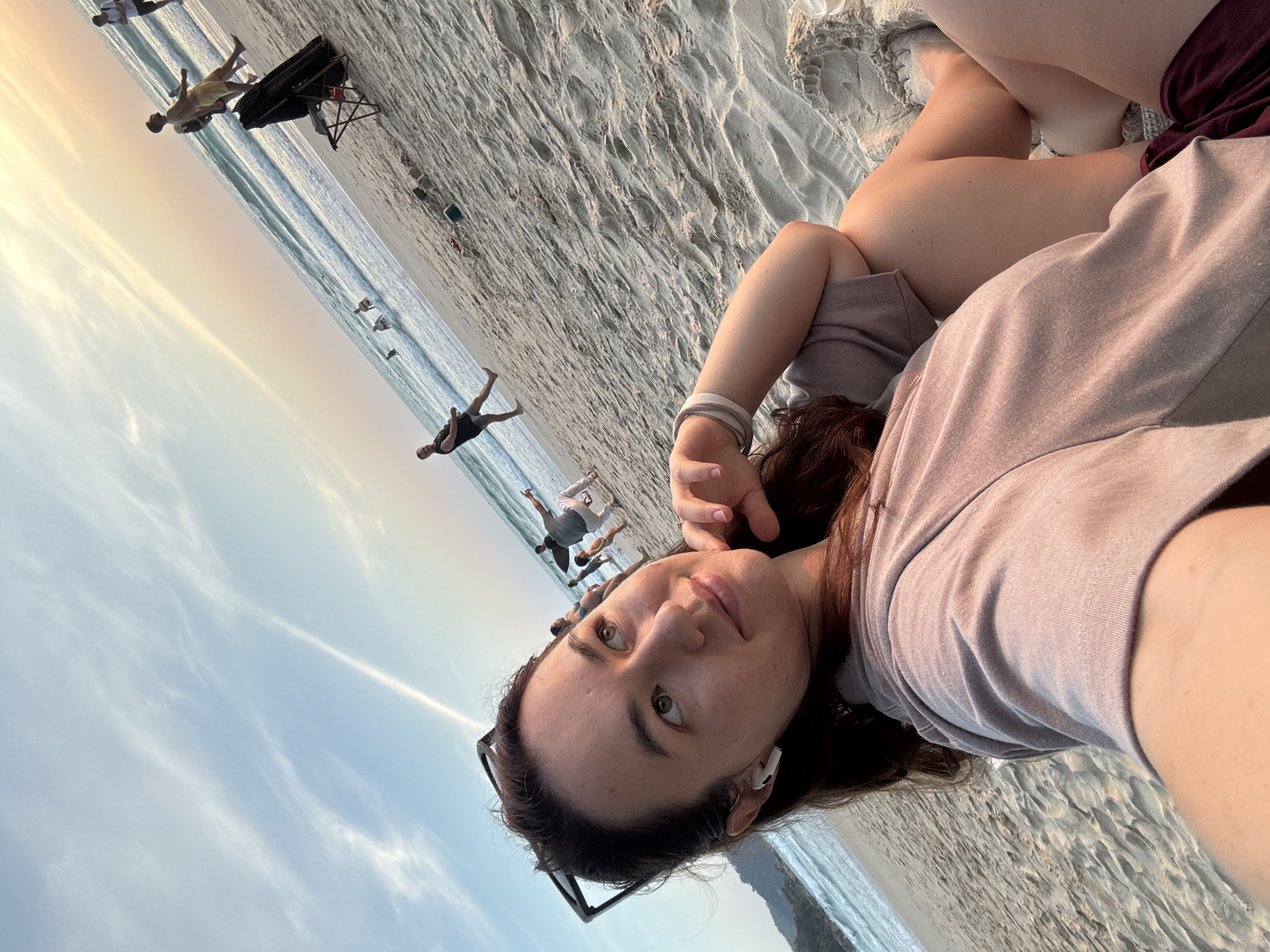 A woman is captured in a selfie at Karon Beach, Thailand, during sunset. She is wearing a grey t-shirt and maroon shorts, with sunglasses perched on her head and an earbud in her left ear. She is reclining on the sandy beach, with her arm resting on her leg. Her gaze is directed upwards and to her right, with a slight smile. In the background, the ocean stretches out, with gentle waves lapping the shore. Several people are visible in the distance, some walking along the beach and others in the water. The sky is a gradient of soft orange and blue, with wispy clouds. A black bag and a folding chair are present on the sand to the right of the frame.