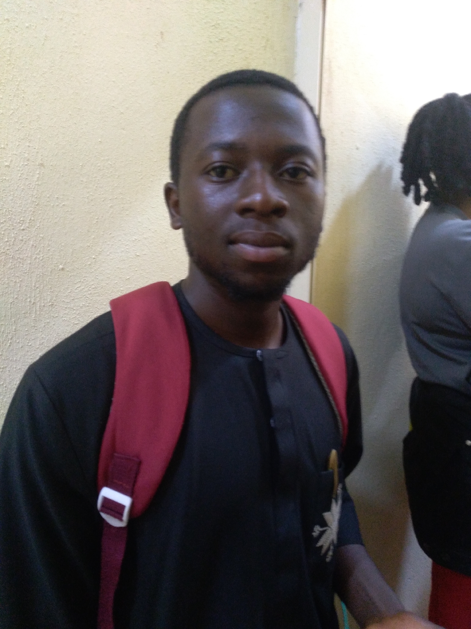 The image portrays a young Black man, likely in Jos, Nigeria, standing indoors and looking directly at the camera. He wears a dark, possibly black or very dark blue, long-sleeved tunic-style shirt with a buttoned placket. A light-colored embroidered design, resembling a stylized plant or star, is visible on the lower right side of his shirt. He carries a maroon or dark red backpack over his left shoulder, with a white plastic buckle prominent on the strap. A yellow or gold pen is tucked into his left breast pocket. The man has short dark hair and a light beard or stubble, and his expression is neutral and composed.
To the right of the main subject, another individual is partially visible from the shoulder up, characterized by dark dreadlocks or braids. The background is a textured, light beige or yellow wall, possibly painted concrete or plaster, with a vertical architectural seam or corner apparent on the right side. The lighting is soft and even, consistent with an indoor setting. The man appears to be posing for the photograph.