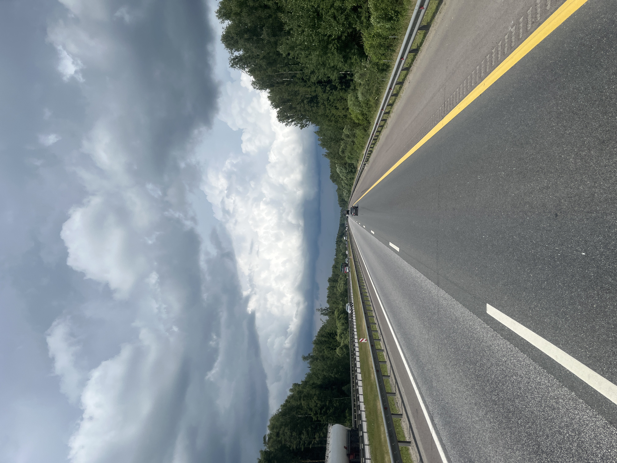 The image depicts a highway in a rural setting, with a dark, cloudy sky overhead. The road has multiple lanes, and there is a guardrail on either side. Dense green trees line the highway. A few cars and a tanker truck are visible on the road. The location in Kiyasovo, Russia, is consistent with a highway traversing a forested area. The scene appears to be captured from a moving vehicle.