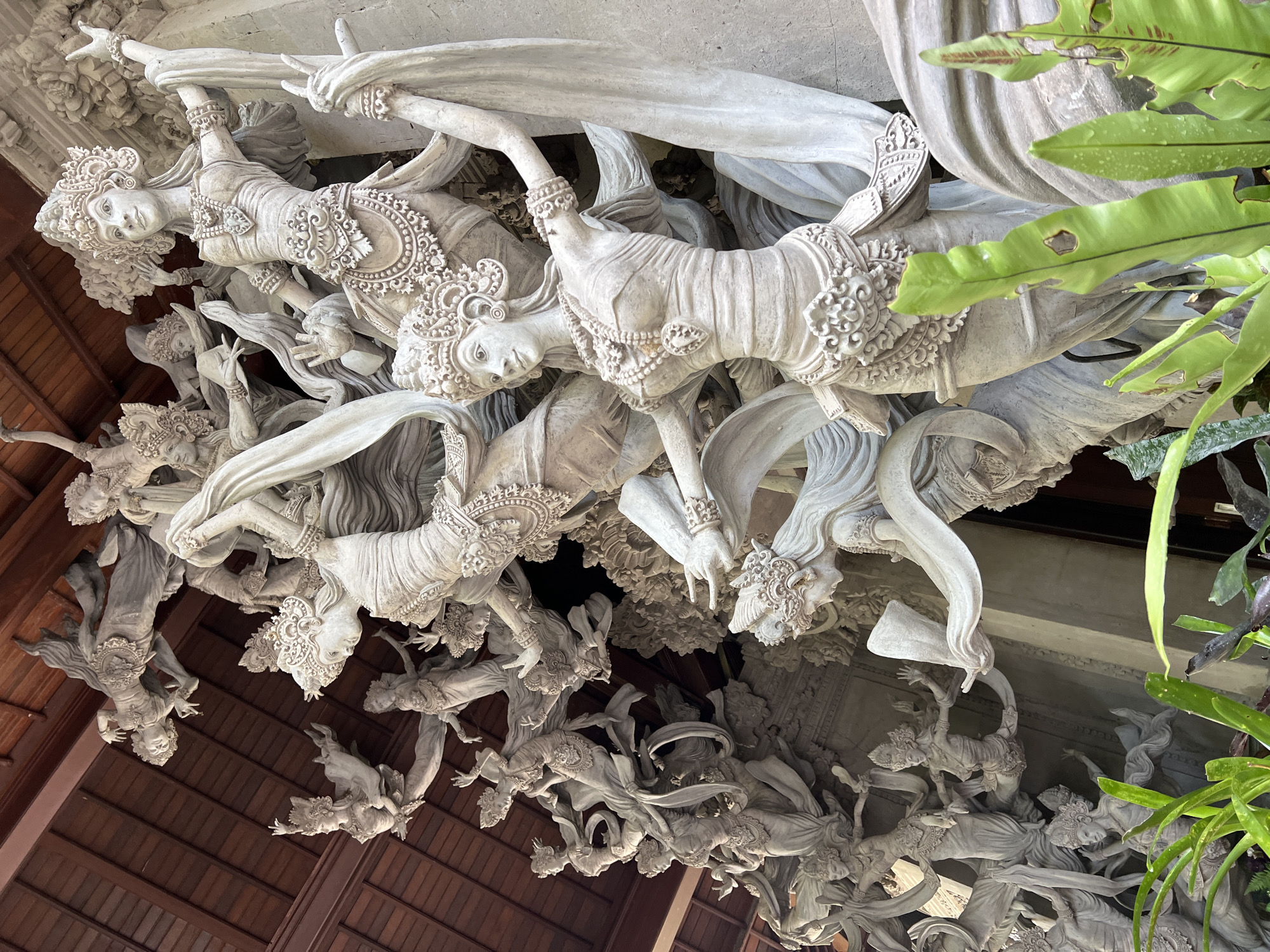 The image depicts an ornate ceiling sculpture located in Kedewatan, Indonesia. The sculpture features multiple figures of dancers carved in white stone or plaster. The figures are adorned with traditional headdresses, jewelry, and flowing garments. They are arranged in a dynamic composition with intertwined limbs and drapery. Portions of a wooden ceiling are visible in the background, along with some green foliage, indicating an outdoor or semi-outdoor setting.