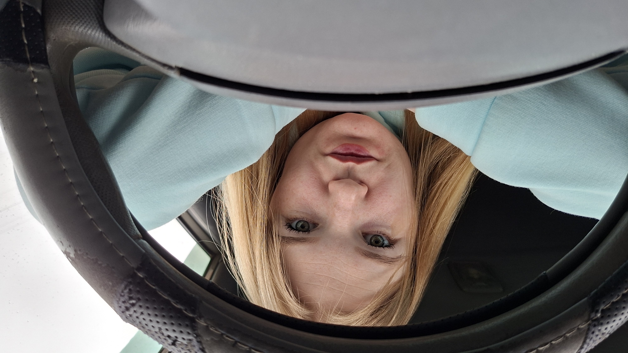 A person with blonde hair and green eyes is visible within the confines of an automobile interior. The individual's face, framed by a light blue garment, is positioned above a steering wheel. The location is not identifiable as Большой Букор, Russia, from the visual data.